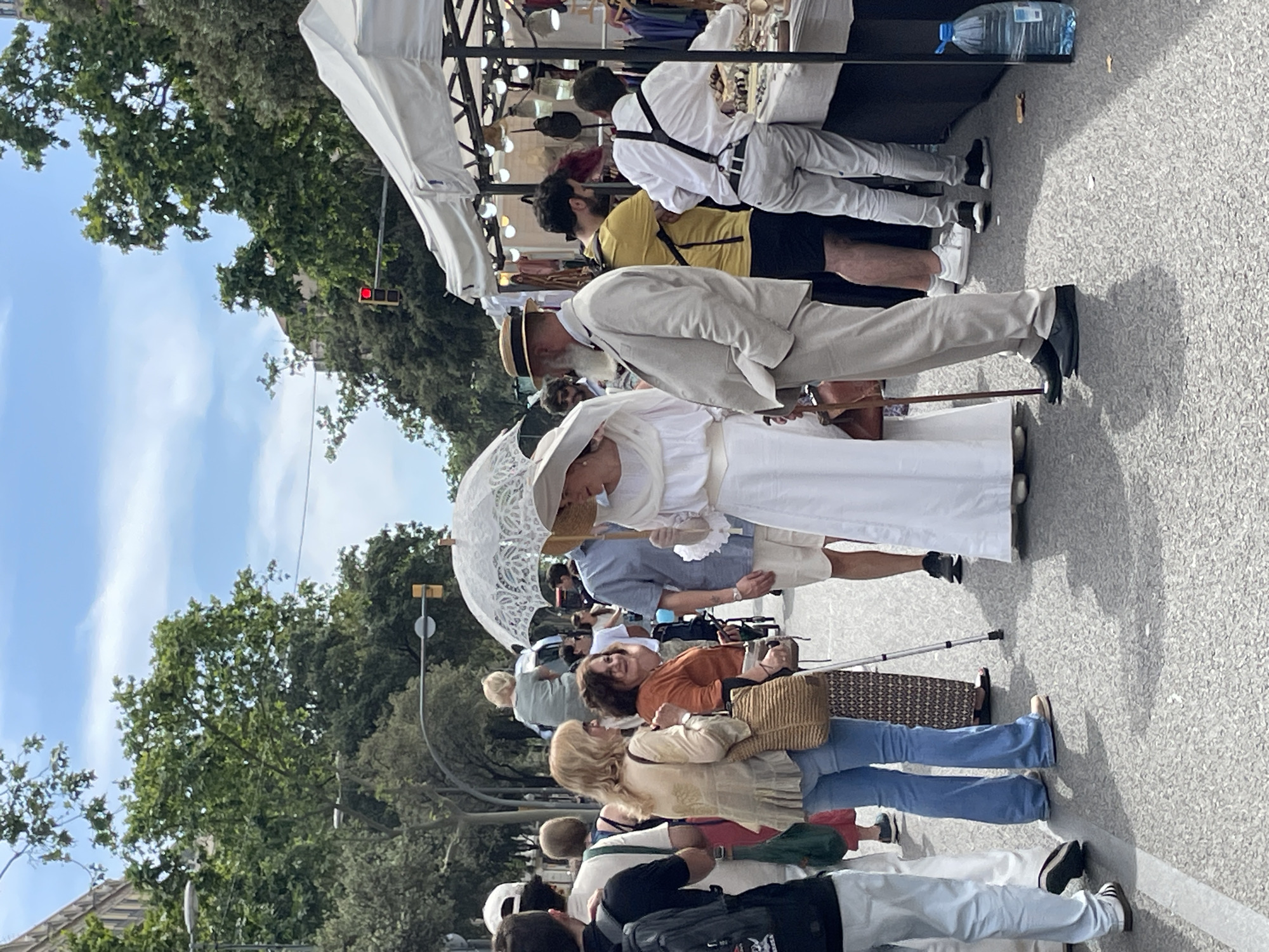 The image shows a street scene in Barcelona, Spain, with a group of people observing a couple dressed in vintage clothing. The couple, a man in a light-colored suit and straw hat and a woman in a white dress with a lace parasol, stand out in the crowd. They are positioned near a market stall with people examining goods. Behind the crowd are trees and buildings, with a traffic light visible in the background. The people in the crowd are wearing casual clothing, and some are carrying bags and using walking aids. The ground is paved, and there is a bottle of water on the stall.
