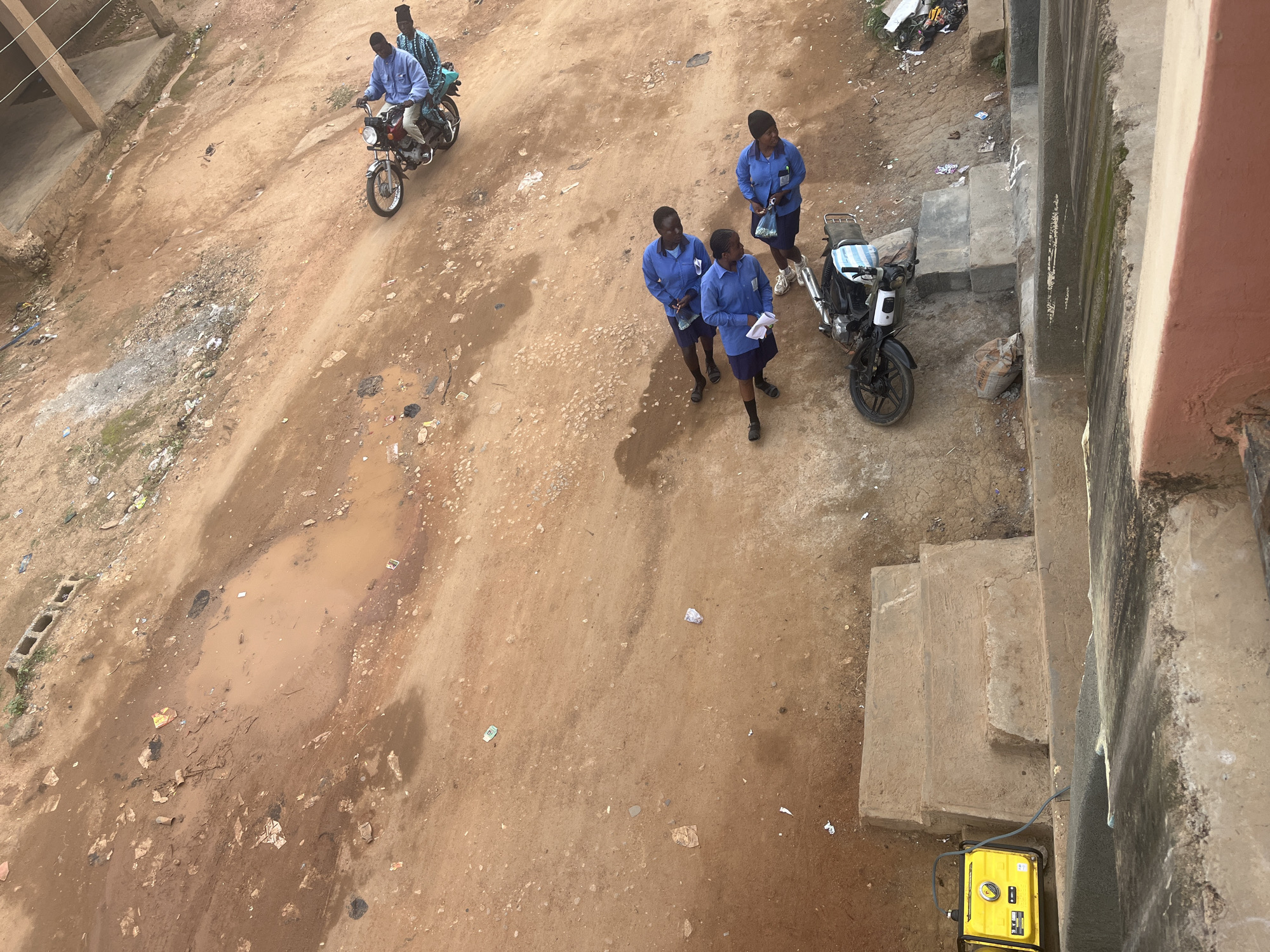In Bukuru, Nigeria, an overhead shot captures a dirt road with a large puddle. Two individuals, dressed in light blue shirts and dark pants, ride a motorcycle. Further down the road, three young people, two appearing to be female and one male, are walking. They are all wearing identical light blue shirts and dark skirts or shorts, suggesting they are students. A motorcycle is parked near them. In the foreground, a yellow generator is positioned on a concrete step.