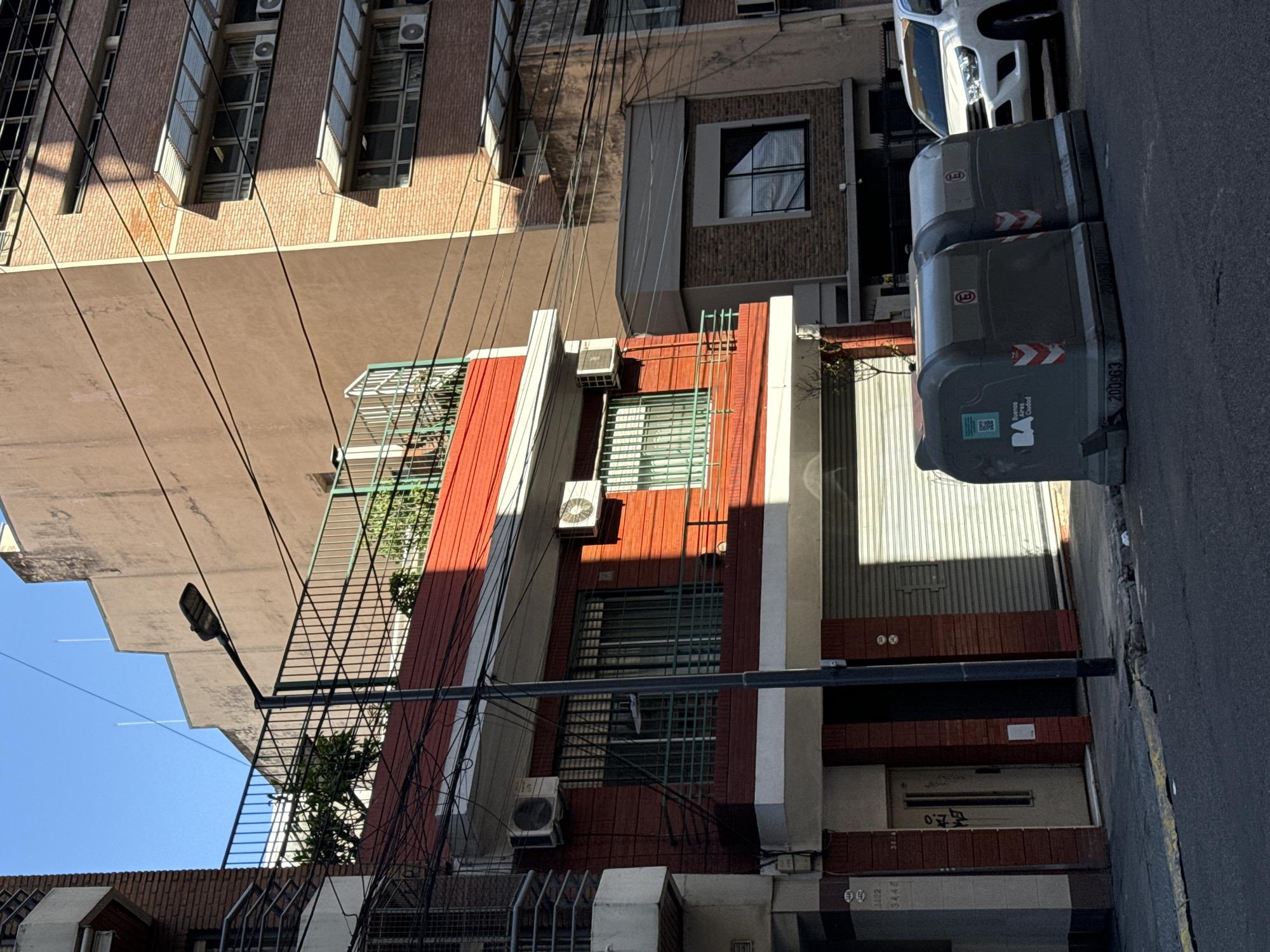 The image captures a street scene in Buenos Aires, Argentina. In the foreground, two gray trash receptacles stand on the right side of the street. Adjacent to them is a sidewalk and buildings of various heights. The architecture includes brick facades, windows with metal grates, and air conditioning units. Power lines crisscross the sky, obscuring parts of the buildings. A street lamp is visible on the left side of the image.