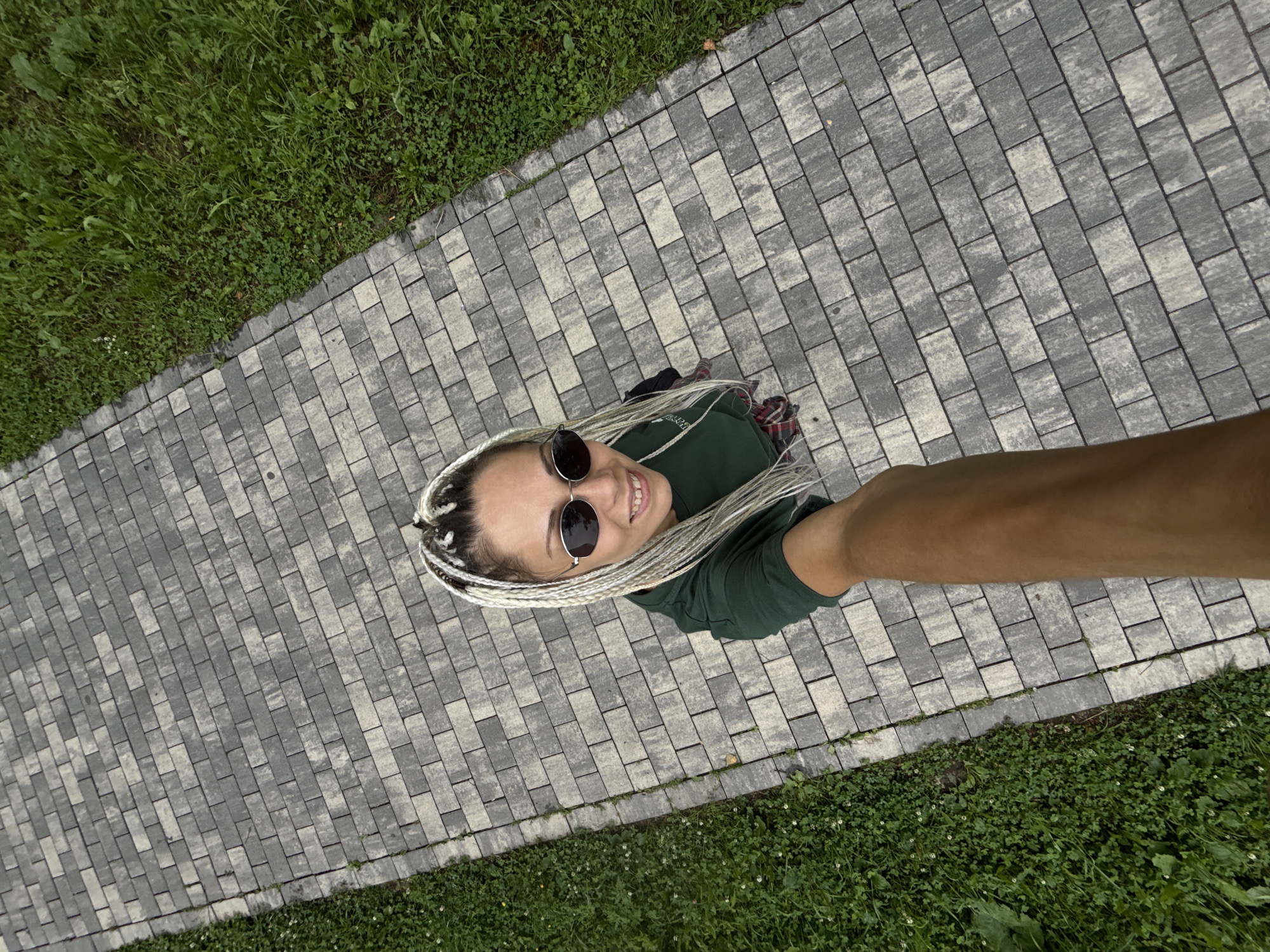 A high-angle, medium shot captures a woman with blonde braided hair and sunglasses smiling as she takes a selfie. She is positioned on a paved walkway composed of rectangular grey stones arranged in a running bond pattern. To the left of the walkway, there is a patch of lush green grass. The woman is wearing a dark green t-shirt and a red and black plaid skirt. Her right arm is extended forward, holding the camera. The image appears to be taken outdoors, with the walkway and grass suggesting a park or garden setting. The specific location in the city of Dumino, Russia, is not discernible from the visual information provided.