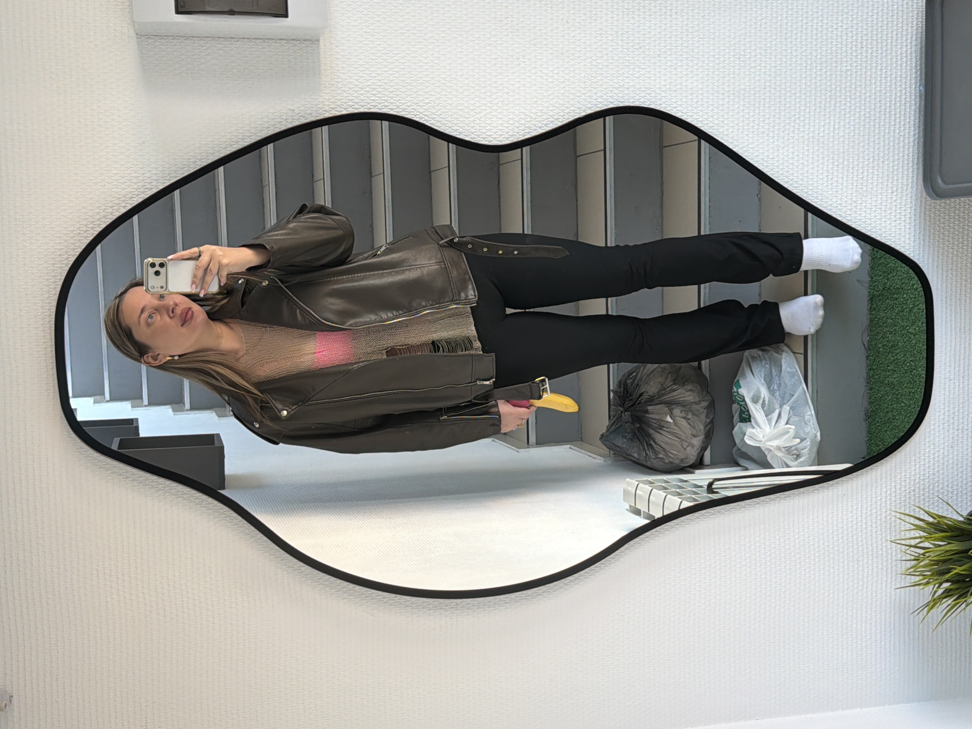 A woman takes a mirror selfie in Chaikovsky, Russia. The mirror is shaped like a pair of lips. The woman is wearing black pants, white socks, a brown leather jacket, and a pink top. She is holding a banana in her left hand and a smartphone in her right hand. The background includes stairs, a trash can, some greenery, and a white wall.