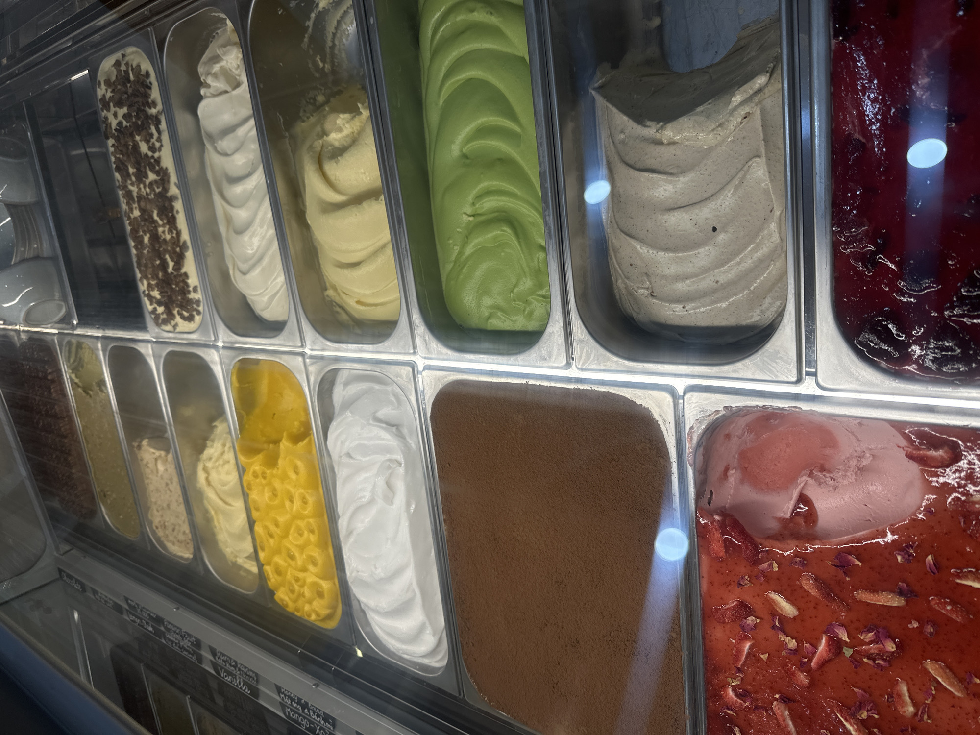 This is a photograph of a gelato display case. The display case is filled with various flavors of gelato, which are contained in metal pans. The gelato flavors include chocolate, mango, vanilla, pistachio, strawberry, and what appears to be a dark berry flavor. The display case is illuminated, and there are reflections on the glass surface. The overall impression is of a well-stocked gelato shop. This scene is likely located in Nha Trang, Vietnam, a popular tourist destination known for its beaches and numerous food establishments.