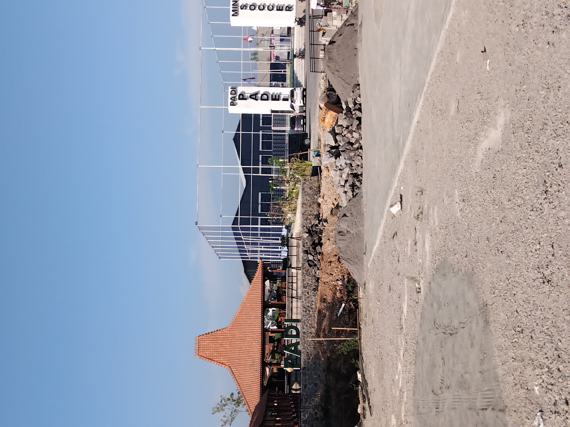 In Papahan, Indonesia, the image captures an outdoor scene featuring a traditional Javanese-style pavilion with a terracotta roof, a mini soccer field enclosed by nets, and a building with dark blue siding and white trim. In the foreground, piles of sand and construction materials suggest ongoing development or maintenance. A prominent sign displays the word "PADI" in large white letters. Two cars are parked near the building, and a motorcycle is visible further in the distance. The sky is clear and blue.