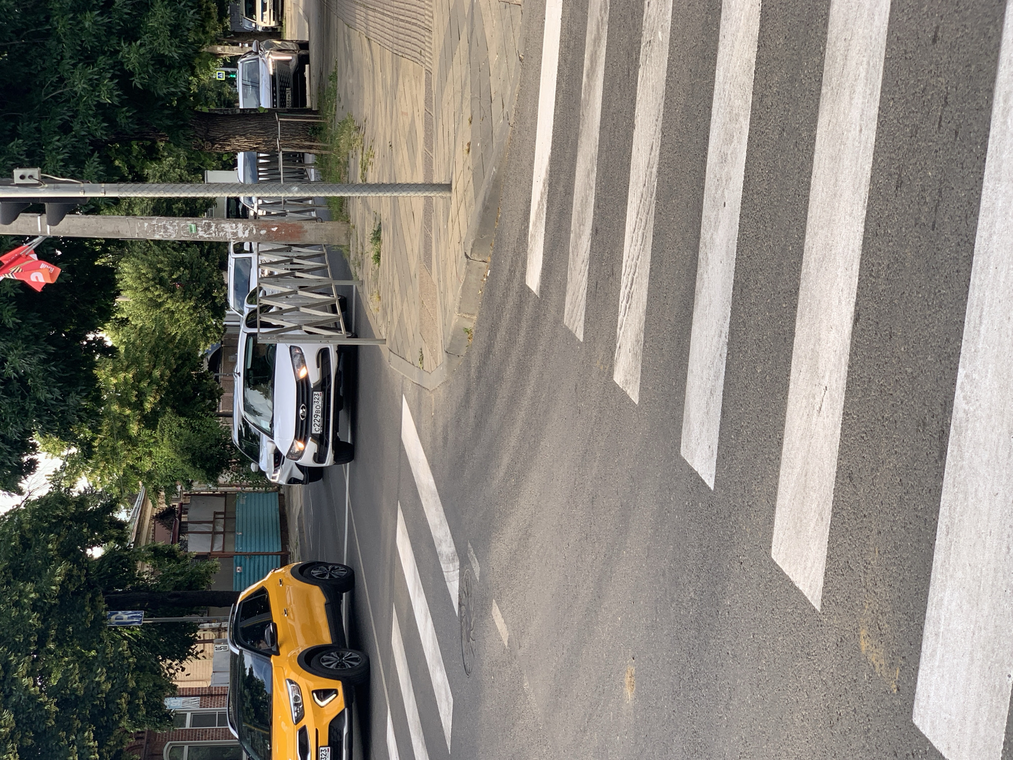 In an urban setting, a yellow Kia SUV and a white Lada sedan are visible at a crosswalk. The yellow SUV is positioned on the left side of the frame, partially obscuring the view of the white sedan behind it. Both vehicles are stopped, suggesting they are yielding to pedestrians or waiting for traffic signals. The crosswalk itself is clearly marked with white stripes on the asphalt road. In the background, trees line the street, indicating a residential or commercial area. A traffic light pole stands to the right of the frame, with a red and white flag attached to it. The overall scene depicts a typical city intersection with vehicles and pedestrian infrastructure.