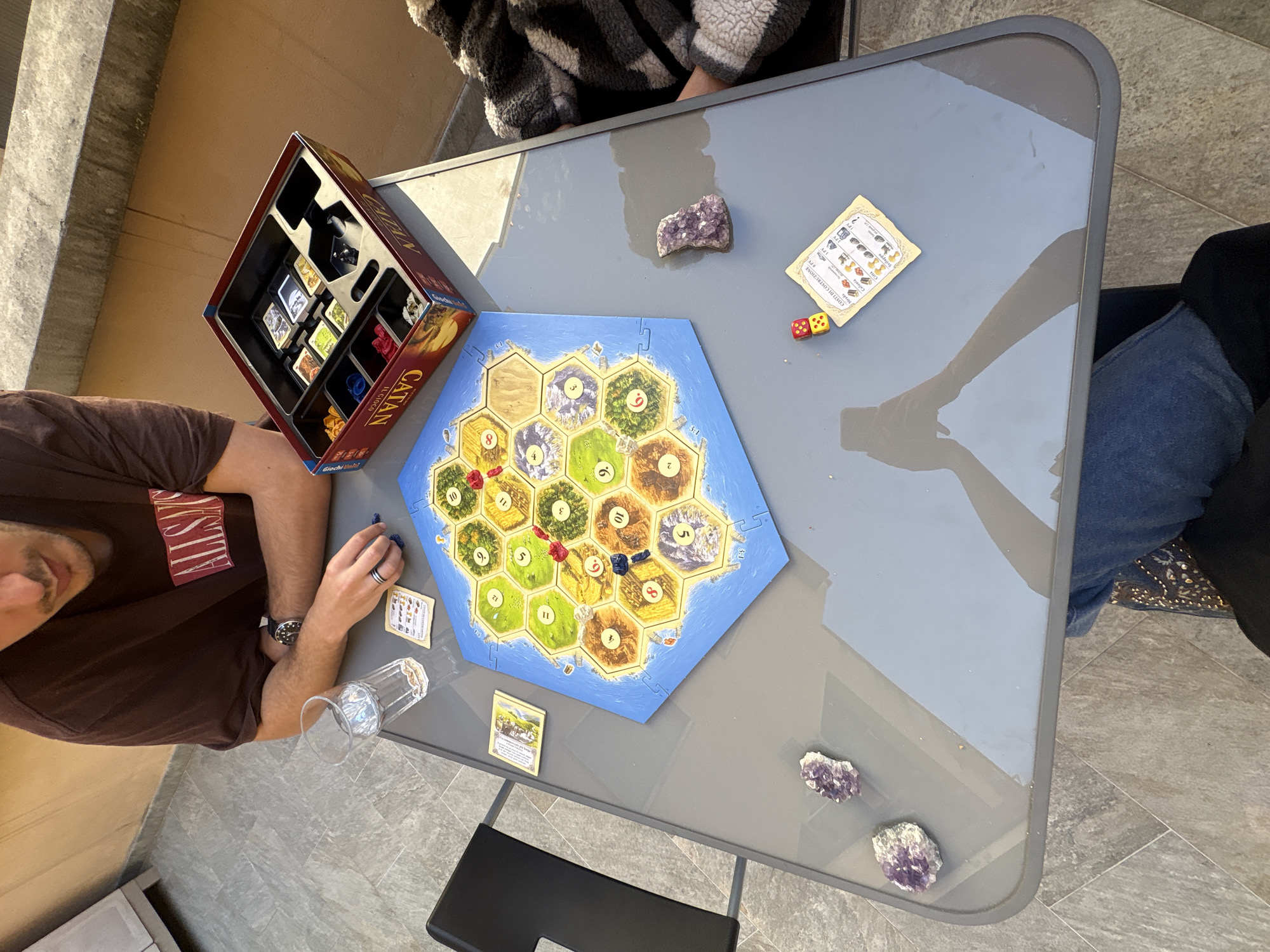 The image shows a game of Catan being played on a gray folding table. A person is seated to the left, wearing a brown t-shirt, with a watch on their left wrist and a ring on their ring finger. Their hand rests near a glass and some cards. The game board is in the center of the table, along with the Catan box and other game components. To the right, someone is wearing blue jeans, and further away another person in a black and white striped jacket can be seen. There are three amethyst crystals on the table around the game. The setting is on a tiled patio, and the image appears to be taken outdoors.