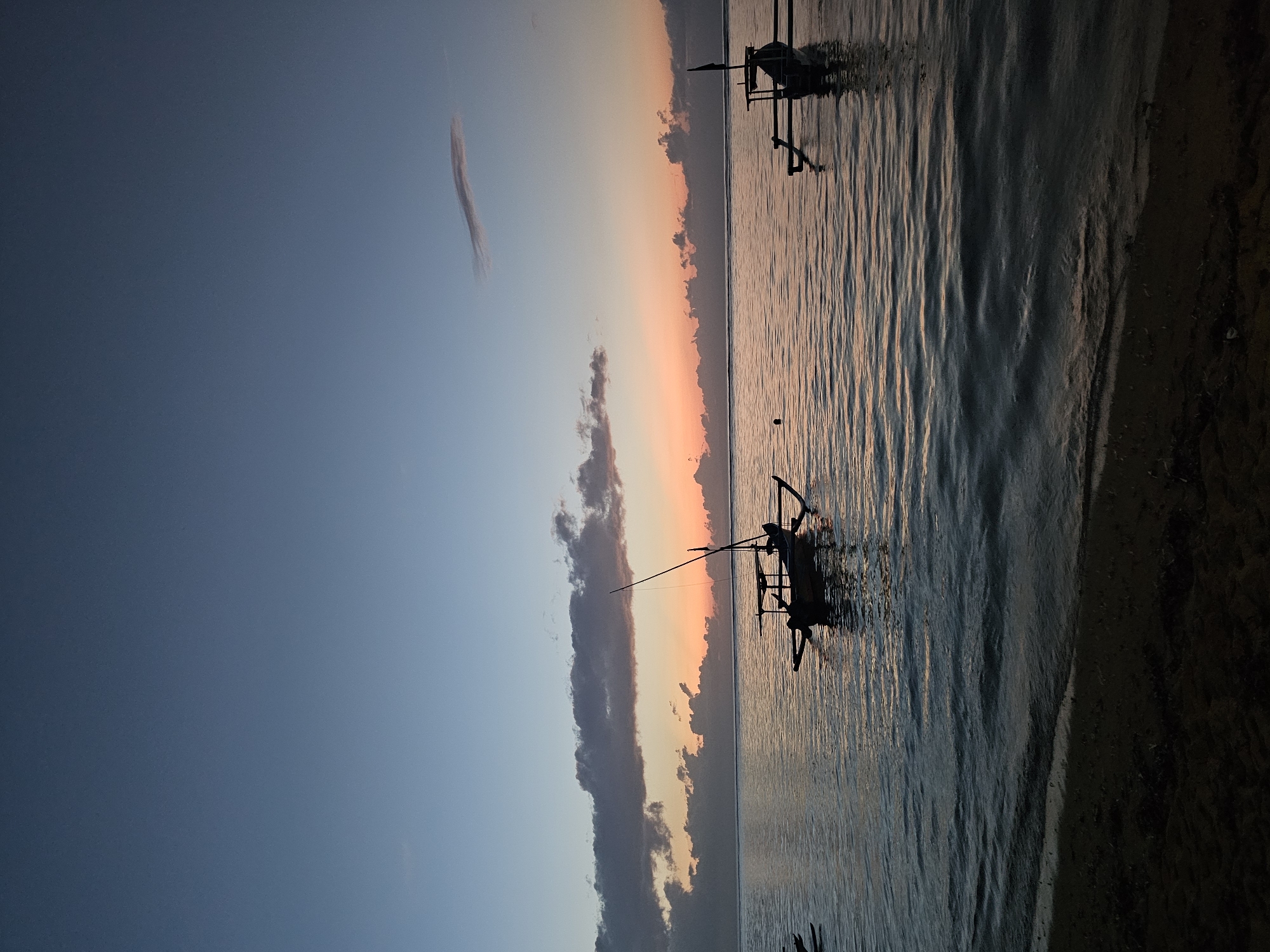 This image depicts a serene seascape at dusk or dawn, likely along the coast of Denpasar, Indonesia. The sky transitions from a deep blue at the top to warm hues of orange and pink near the horizon, indicating the presence of the sun just below the visible edge. Scattered clouds, both dark and wispy, add texture to the sky.
The calm sea reflects the colors of the sky, with gentle ripples on its surface. In the foreground, a sandy shore is visible, with the dark, wet sand suggesting it is close to the waterline.
Dominating the water are two traditional Indonesian outrigger boats, silhouetted against the colorful sky. The boat in the middle ground is closer to the viewer and features a prominent mast. Another similar boat is visible further to the right. The boats appear to be anchored or drifting peacefully. No people are discernible in the image. The overall atmosphere is tranquil and natural.