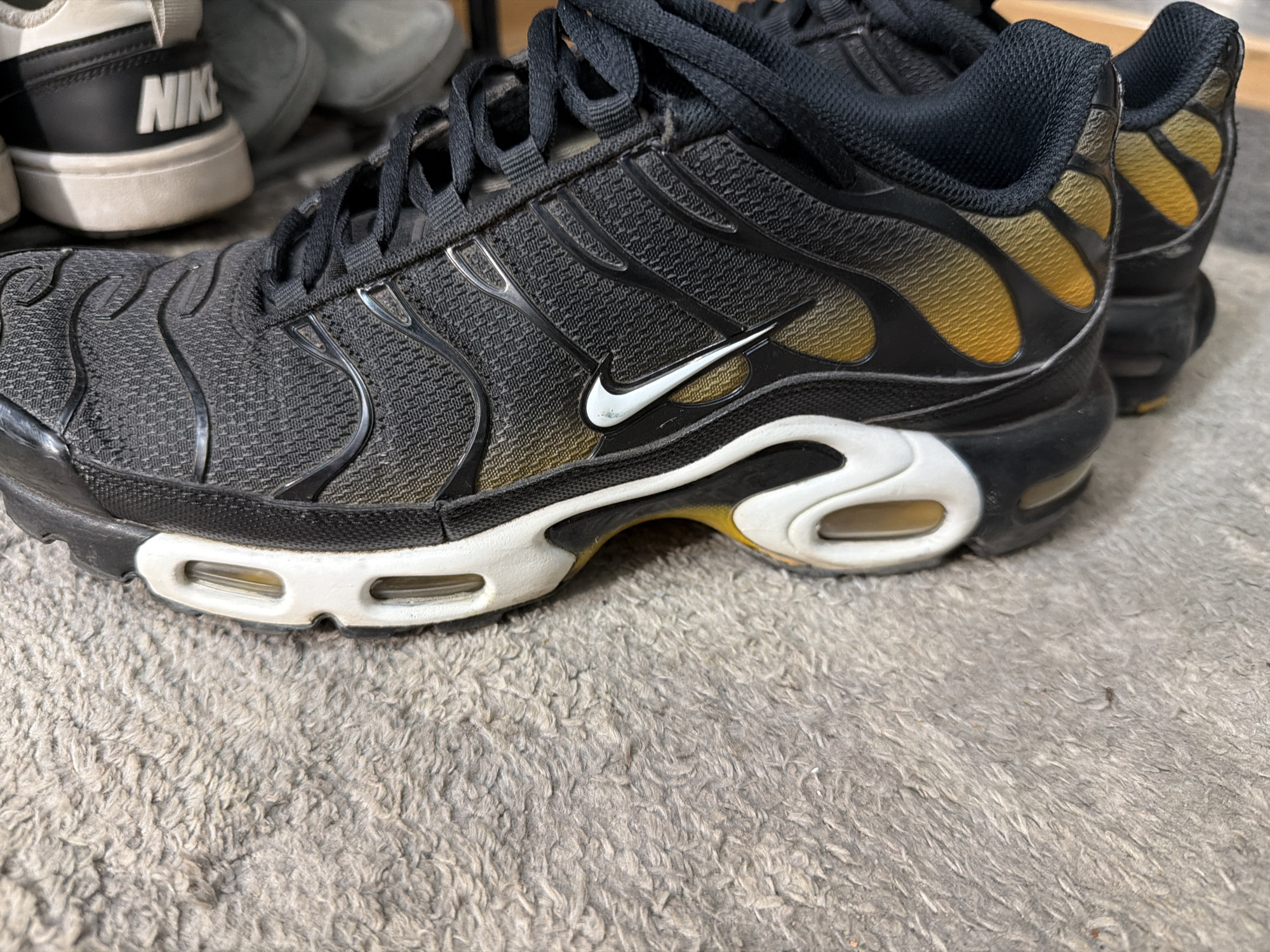The image shows a close-up of a black Nike Air Max Plus sneaker resting on a gray, textured surface, likely carpet. The sneaker features a white Nike swoosh logo on the side, a gradient gold-to-black pattern towards the heel, and a white midsole with visible air cushioning. Another pair of Nike sneakers is visible in the background. The location is not ascertainable from the image.