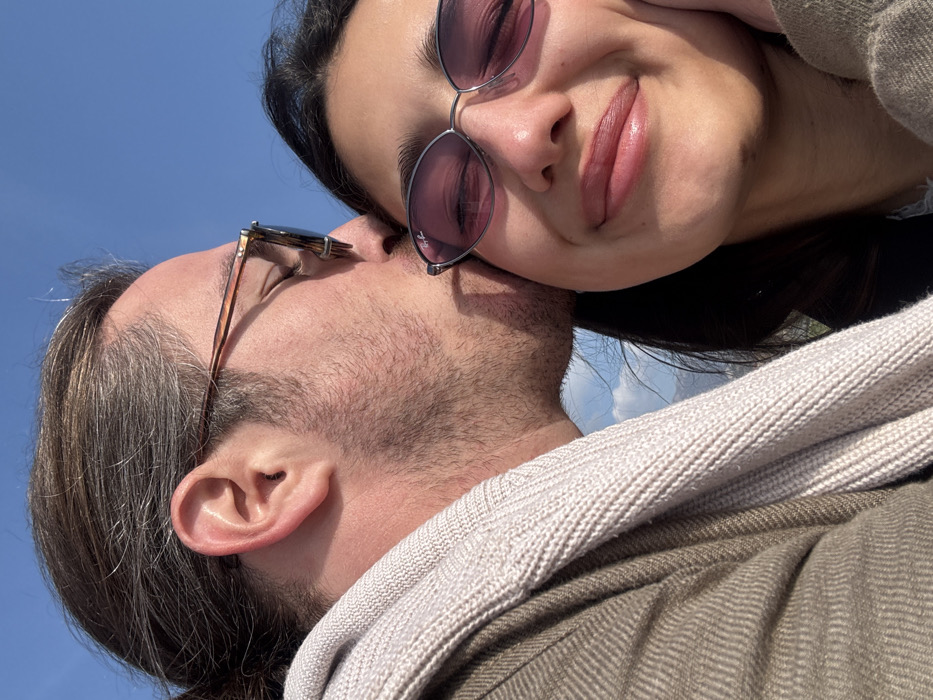 The scene is an outdoor setting in Šušanj, Montenegro, under a clear blue sky with some distant white clouds. Two individuals are depicted in a close-up, chest-up vertical frame. On the left, a male subject with dark hair pulled back, stubble, and tortoise-shell eyeglasses (Ray-Ban branded) is shown in profile, leaning into the female subject. He wears a light beige knitted scarf or sweater over a herringbone-patterned jacket. He is kissing the right cheek of the female subject, who occupies the right side of the frame. The female subject has dark hair, wears silver-framed oval sunglasses with purple-tinted lenses (Ray-Ban branded), and exhibits a slight smile, revealing dimples, with her eyes closed or partially closed. Her right hand's fingers lightly touch her cheek/jawline. Her lips appear full. She wears a dark outer garment over a lighter inner layer.