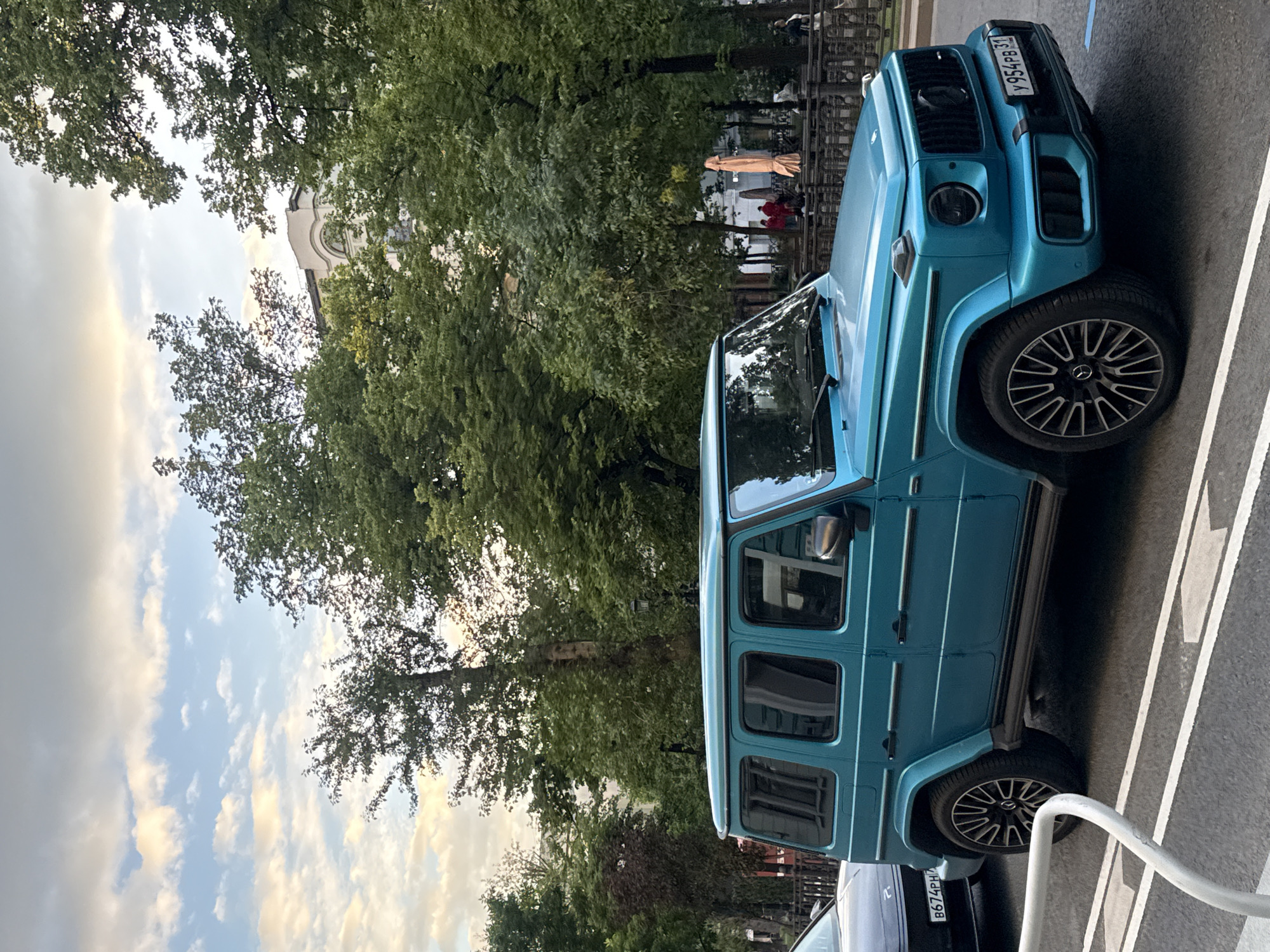 The image, rotated, captures a bright turquoise Mercedes-Benz G-Class SUV (G-Wagen) parked on an asphalt surface, likely a street or parking lot, in an urban setting. Lush green trees dominate the background and frame the sky, which features both blue patches and scattered white clouds, suggesting a pleasant late afternoon or early evening.
The G-Wagen is a prominent subject, showcasing its distinctive grille, headlights, and black multi-spoke alloy wheels. Its front license plate, visible at the top right, displays the Russian registration "Y954PB31." A portion of another vehicle, including its rear license plate "B674PH," is visible behind the G-Wagen. White arrow markings and parking lines are painted on the asphalt, and a white metal barrier is in the foreground.
In the mid-ground, beyond the trees, faint figures of people can be discerned. One individual appears to be wearing a long, light-colored robe or garment, while a small group in the distance wears red. A section of a light-colored building with an arched window, possibly a historic structure or church, is partially visible through the foliage. The scene depicts a quiet, parked street in what is contextually identified as Moscow, Russia.