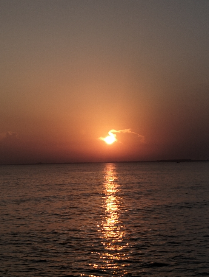 An orange sun sits on the horizon, partially obscured by a cloud. A brilliant reflection of the sun stretches across the water towards the viewer. The sky transitions from a light orange near the horizon to a dusky grey at the top of the frame. A distant landmass is visible as a dark line along the horizon. The water is rippled, catching the sun's rays.