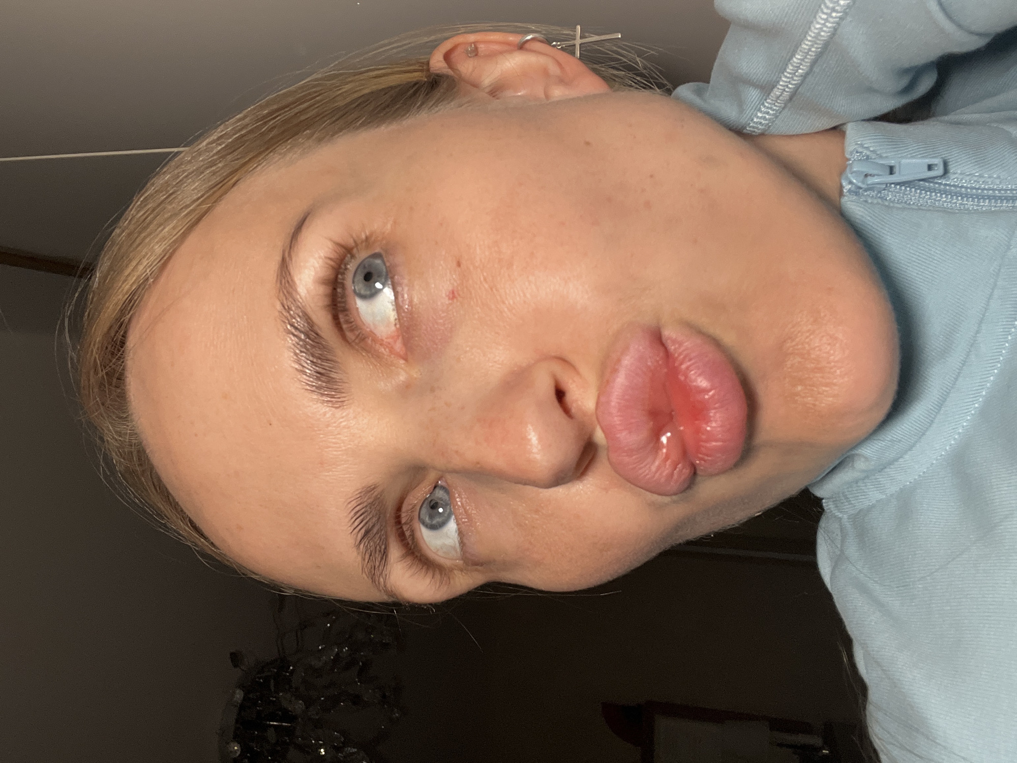 A close-up, slightly angled, medium shot shows a young Caucasian woman with blonde hair, pale skin, and blue eyes. Her lips are pursed in a pout, and she is looking up and to her right. She is wearing a light blue zip-up hoodie. An earring with a cross pendant is visible on her right ear. The background is dark and out of focus, with a glimpse of a chandelier on the left and a plain grey wall on the right. The lighting appears to be artificial and casts shadows on her face. The image does not provide any information about the location being in a city in Russia.