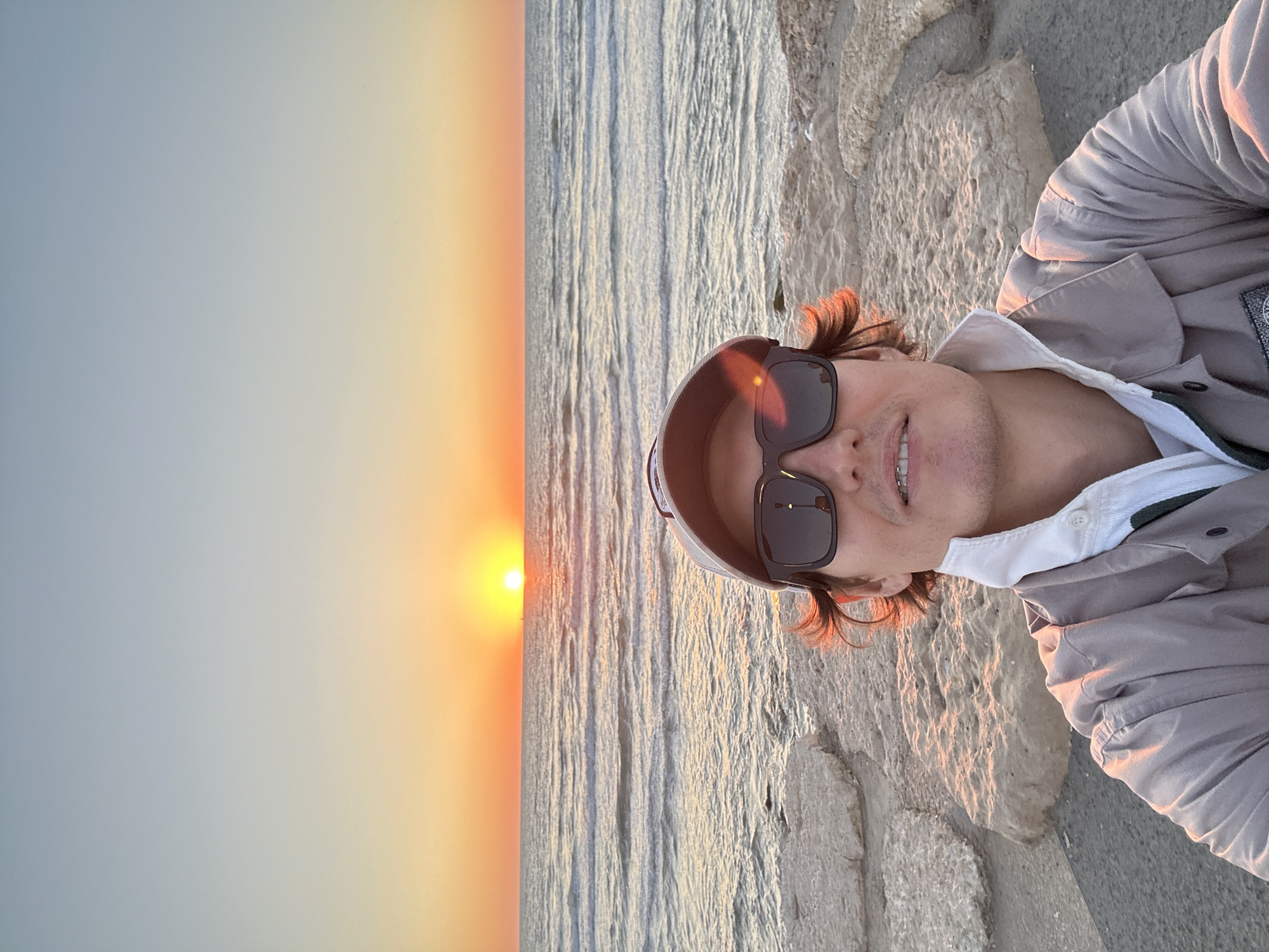 The image captures a man taking a selfie or being photographed at a coastal location during sunset or sunrise. The man is positioned on the right side of the frame, smiling slightly towards the camera. He has light brown, wavy hair and wears dark sunglasses, which reflect the bright sun. He is dressed in a light grey or beige long-sleeved jacket over a white collared shirt, with a hint of a darker collar or undershirt visible. A circular badge, identifiable as the "Stone Island" compass logo, is clearly visible on the left arm of his jacket.
To the left, the vibrant orange sun is low on the horizon, casting a warm glow across the entire scene. The sky transitions from a soft blue-grey towards the top to rich orange and yellow hues near the sun. Below the horizon, a body of water, likely the sea or ocean, stretches across the middle of the frame, its surface reflecting the light with numerous ripples and gentle waves. On the far right, behind the man, there's a textured, light-colored rocky or concrete embankment or wall. The clear weather and warm, golden hour lighting contribute to a serene and pleasant atmosphere, characteristic of a coastal setting like Tel-Aviv, Israel.
