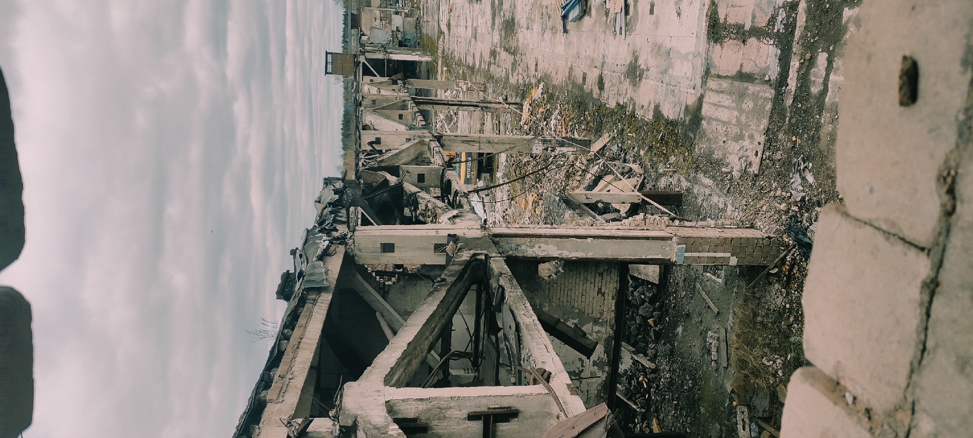 The image depicts the ruins of a building, likely in an industrial area of Fryazino, Russia, judging by the architectural style. The structure is heavily damaged, with exposed concrete and brickwork, collapsed beams, and scattered debris. The sky is overcast, casting a dull light on the scene. There is no evidence of human presence.