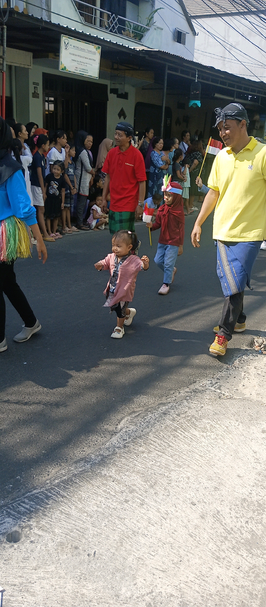 In Malang, Indonesia, on a sunny day, a lively scene unfolds with a parade or procession. The foreground is dominated by a young child, possibly a girl, in a pink outfit, with dark hair and bare legs, actively walking or running on an asphalt road. Beside her, slightly further back, a child in a maroon outfit and headscarf, holding a small Indonesian flag on a stick, is also walking. To the right, an adult man wearing a yellow polo shirt and a blue sarong walks past, holding another Indonesian flag.
In the background, a diverse crowd of adults and children are gathered, lining the street and observing the event. Some are standing, while others appear to be sitting or squatting. Several individuals are wearing traditional Indonesian attire, including headscarves and sarongs. A red banner with text in Indonesian hangs on a building on the left side of the frame, indicating a business or organization named "PT. MIONS CAKRA TEKNOLOGI". The environment suggests a street in a residential or commercial area, with buildings and some overhead wires visible. The overall atmosphere appears festive and community-oriented.