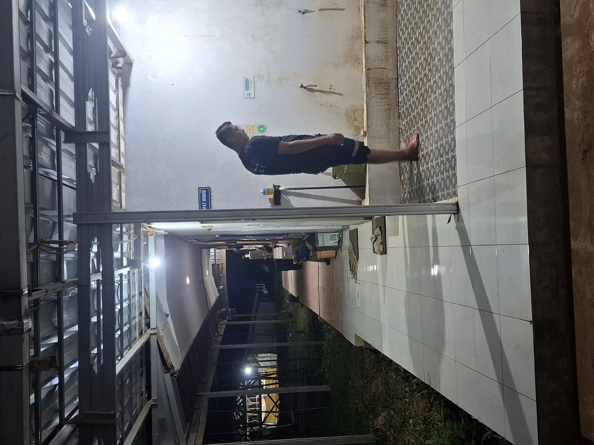 A man stands facing a white wall with faucets, in what appears to be a public ablution area. He is wearing a dark t-shirt and shorts. The area is tiled and there are a few buckets and mats visible. To the left, a walkway with white tiles leads into a dimly lit covered area with metal supports. The scene is outdoors, possibly at night, with artificial lighting. A sign above the man reads "TEMPAT WUDHU," indicating it is a place for ritual washing. The overall environment suggests a facility likely located in a place like Kutasirna, Indonesia, given the contextual information provided.