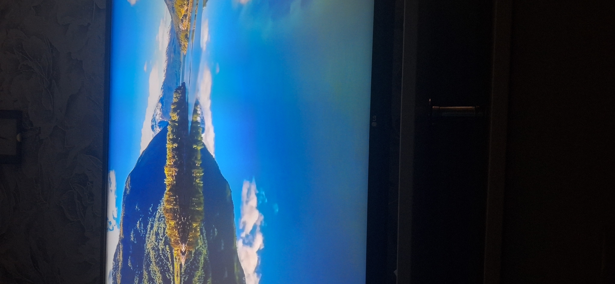 The image displays a scenic view of a body of water reflecting mountains and trees under a clear blue sky. In the foreground, the lower part of a television screen is visible, showing a black frame with an "LG" logo on the right side. The background of the image, above the television screen, appears to be a dark, textured wall. There is no discernible information within the image to suggest a location in Bila Tserkva, Ukraine, or any specific urban setting. The scene is a natural landscape.