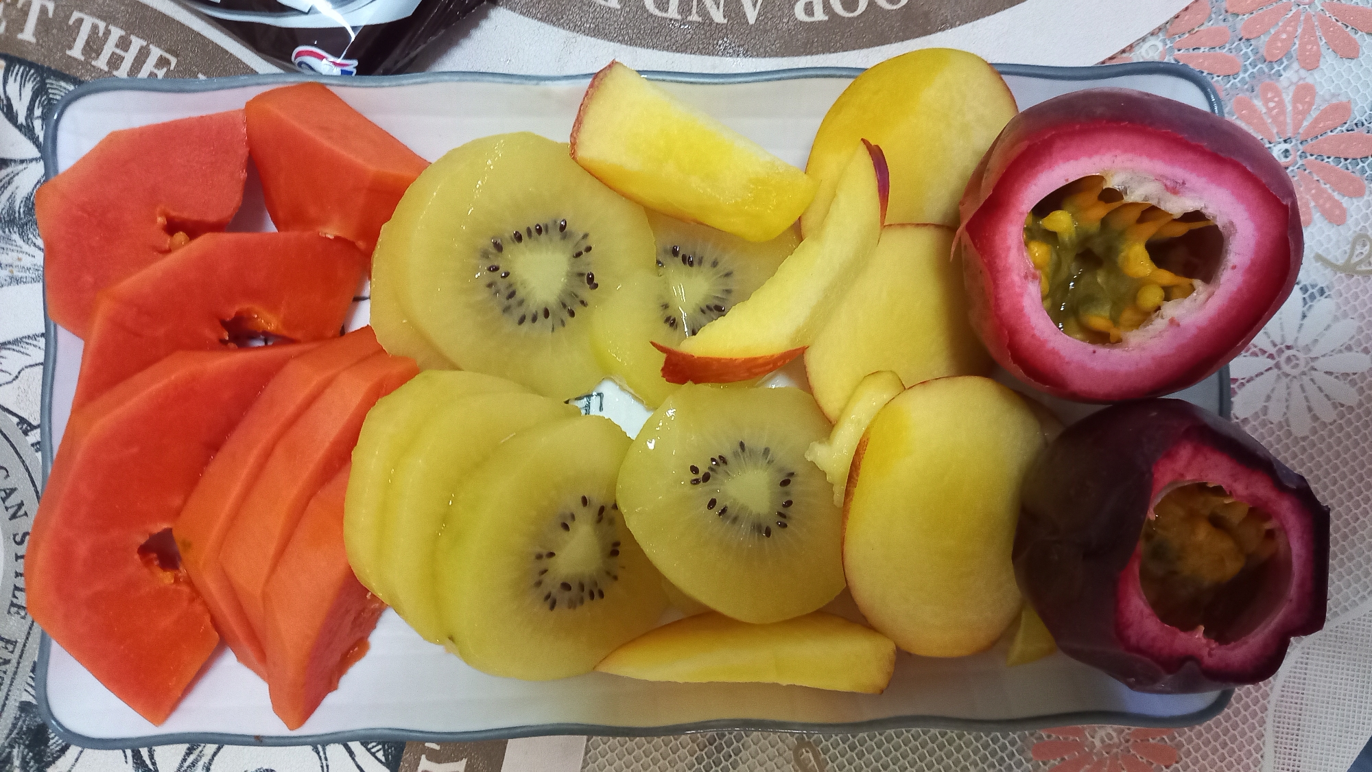 A close-up, overhead shot displays a white rectangular plate filled with a colorful assortment of sliced fruits. The plate is positioned on a surface adorned with a lace-like tablecloth featuring floral patterns in pink and white. The fruits are arranged artfully, creating a visually appealing presentation. Dominating the top portion of the plate are slices of bright orange papaya, cut into irregular wedges. Beneath the papaya, a layer of golden kiwi slices is visible, their characteristic black seeds forming a concentric pattern within the vibrant yellow flesh. Interspersed with the kiwi are slices of light yellow fruit, possibly peaches or nectarines, some with a hint of red blush on their skin. In the foreground, two halves of a passion fruit are prominently displayed. One half is a deep reddish-purple, while the other exhibits a lighter pinkish-purple rind. Both passion fruit halves reveal the juicy, gelatinous pulp and seeds within, in shades of yellow and green. The overall impression is one of freshness and a refreshing, tropical treat. The background shows a textured surface with printed text in English, including phrases like "CAN STYLE ENT" and "GOOD AND," suggesting a possibly commercial or decorative context, although the specific location within Tambon Koh Phangan, Thailand, cannot be determined from the visual evidence alone.