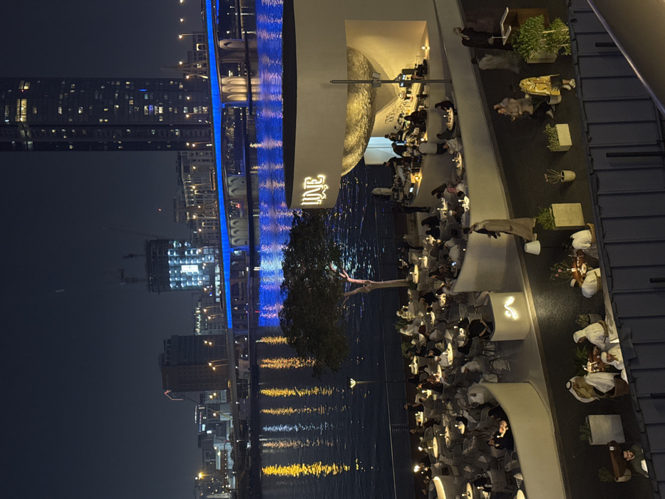 An outdoor restaurant or lounge sits next to a body of water, likely a river or bay. The establishment is bustling with patrons seated at numerous tables. Behind the restaurant, a cityscape is visible, featuring illuminated buildings and a bridge with blue lights. Reflections of the city lights shimmer on the water's surface, creating a vibrant night scene.