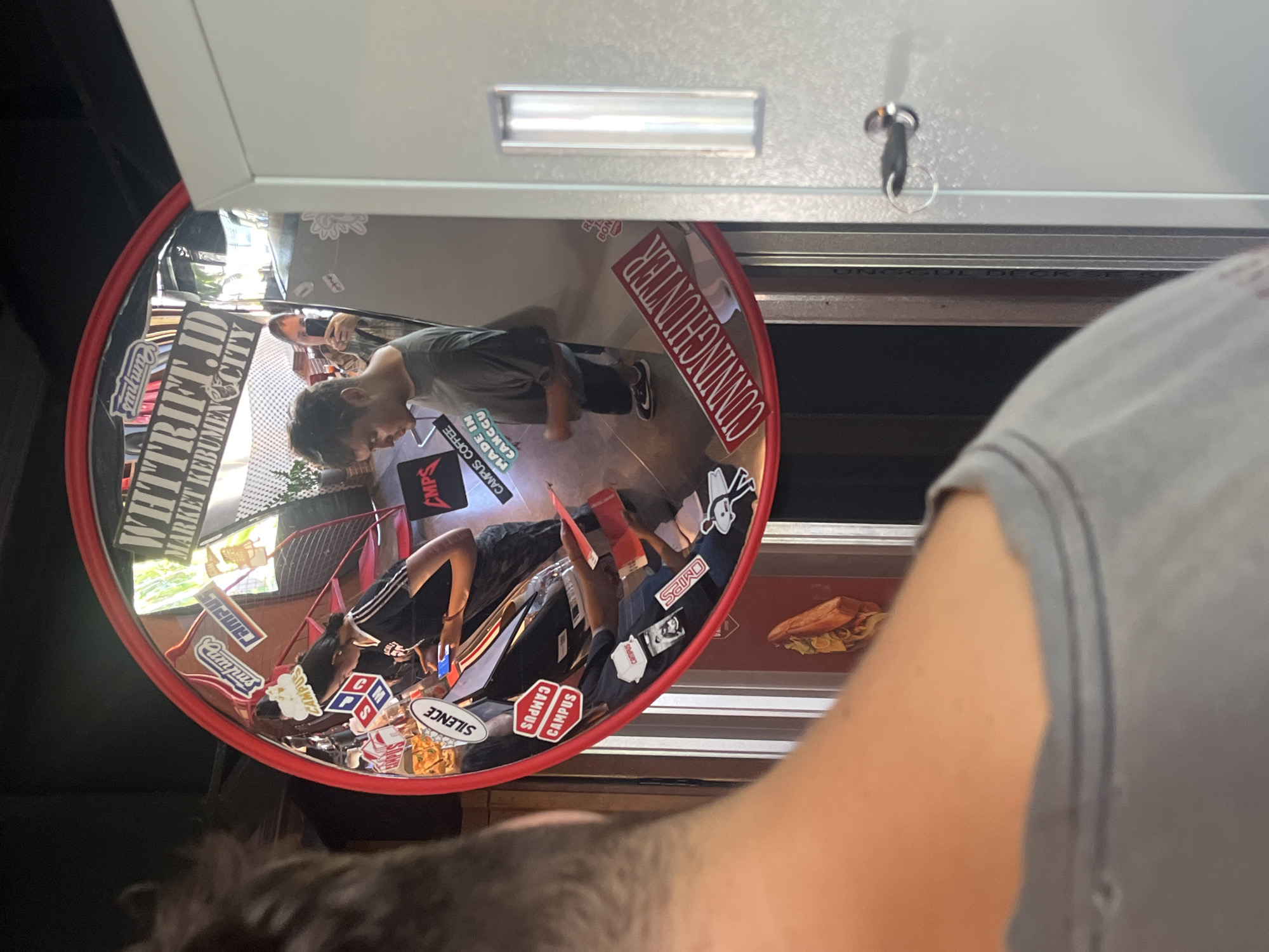 A round mirror, decorated with stickers, reflects a café's interior. Stickers such as "WHTTRIFT.ID MARKET KEBUMEN CITY," "CAMPUS COFFEE MADE IN CANGGU," and "CUNNINGHUNTER" are affixed to the mirror's frame. Reflected within the mirror is a café scene with a person wearing a grey shirt looking at their phone, another person in a black tank top and black and white athletic pants, and a partially visible red basketball hoop. The café appears to be located in North Kuta, Indonesia. The mirror is mounted on a white cabinet with a key hanging from it. A person with grey hair and a grey shirt partially obscures the view of the mirror.