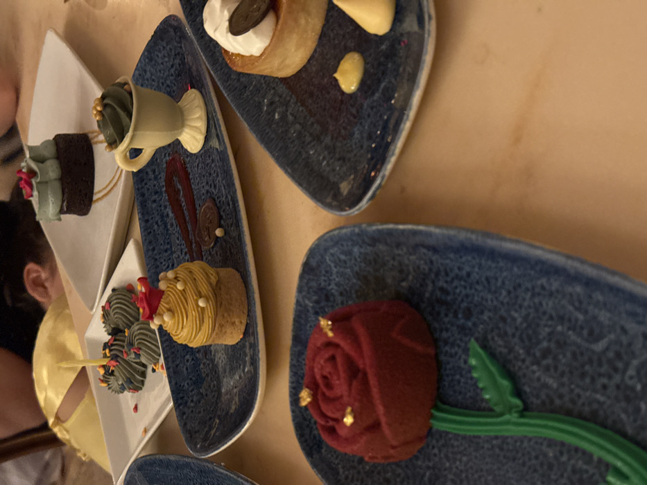 The scene depicts a selection of themed desserts arranged on a light brown dining table within a restaurant setting. This location is identified as Be Our Guest Restaurant in Fantasyland, Magic Kingdom Park, Walt Disney World Resort, Bay Lake, Florida, United States.
In the foreground, to the lower left, a dark blue speckled rectangular plate holds a red, rose-shaped dessert with a textured, matte finish, adorned with small gold flakes on its petals. A sculpted green stem and leaf extend from its base onto the plate.
Adjacent and slightly further back, another dark blue speckled rectangular plate displays two primary desserts: a circular pastry base topped with a tall, swirling mound of mustard-yellow frosting decorated with small white spherical candies and a tiny red flower at its apex, and a white, molded ceramic cup resembling a teacup, containing dark olive-green frosting topped with gold sprinkles. Between these two items, a dark red berry coulis is artfully drizzled in a zigzag pattern, accompanied by small white and dark brown spherical garnishes.
To the upper right, a third dark blue speckled plate is partially visible, featuring a round tart or cheesecake topped with white whipped cream and a flat, dark circular chocolate garnish. Two small, pale yellow dollops of sauce are present on this plate.
Further into the background, on a white rectangular plate, multiple small, dark grey-green swirled frosting portions, likely on mini bases, are visible, some scattered with colorful sprinkles. A thin, lit yellow candle stands upright among these. Directly behind this, on a white plate, sits a small, dark chocolate brownie topped with light mint-green frosting and small pink rosette details.
In the blurred background, partially visible, are the upper torso and head of an individual wearing a yellow garment, possibly a character costume or a guest's attire. Another person's ear is also indistinctly visible. The overall impression is a festive dessert service, possibly celebrating an event given the presence of a lit candle.