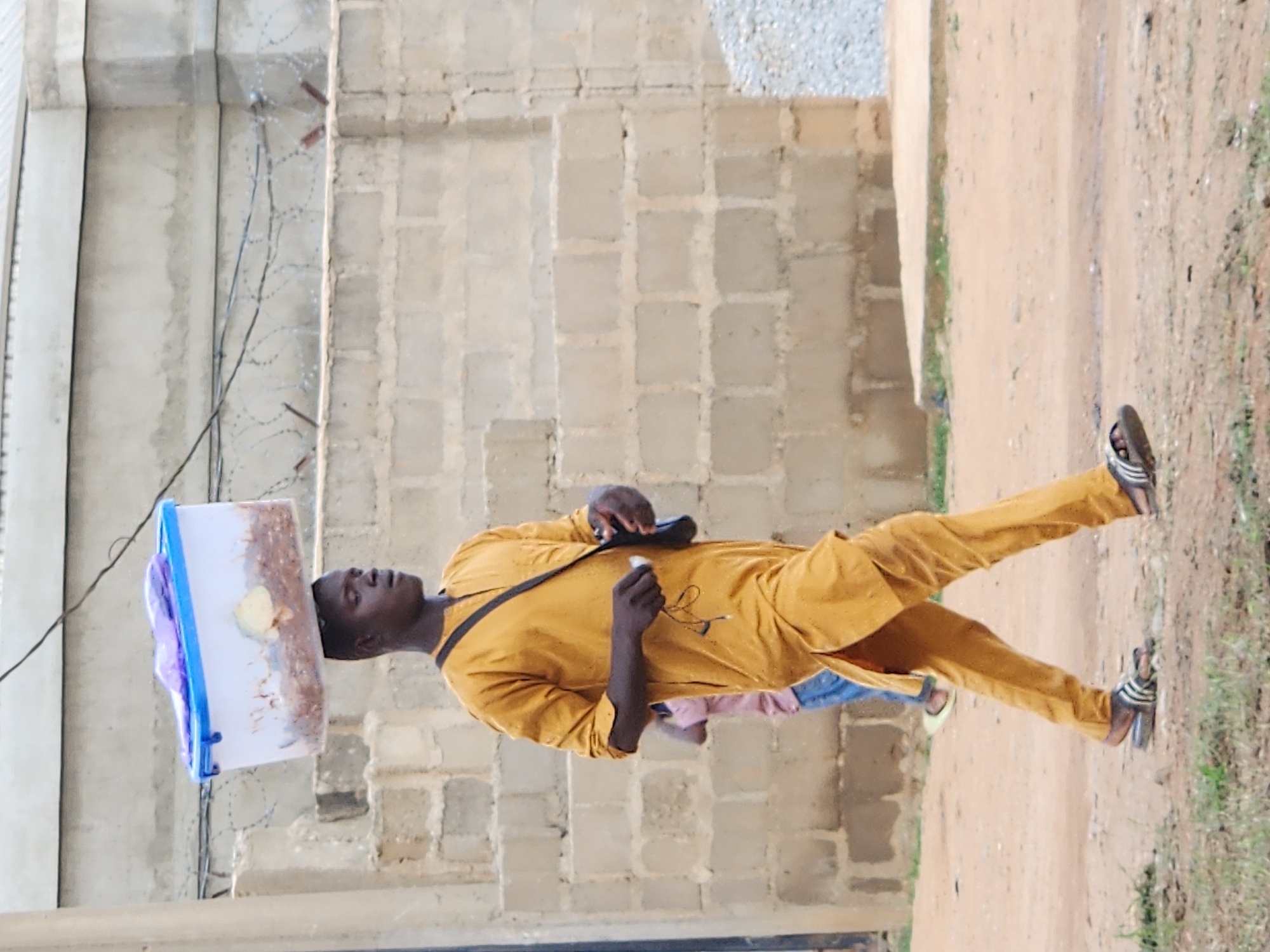 A young Black man walks from right to left in the frame, carrying a white plastic container with a blue rim on his head. The container appears to hold a light-colored, textured substance, possibly food, covered by a purple plastic bag. He is wearing a mustard-colored, long-sleeved tunic and pants. A dark bag is slung over his right shoulder, and he holds what appear to be white earbuds and a wire in his left hand. He is wearing striped flip-flops. In the background, another person, appearing younger and wearing a pink shirt and blue pants, walks away from the viewer. The ground is dirt with patches of dry grass. Behind them is a wall constructed of light-colored cinder blocks. A wire fence with barbed wire runs along the top of a portion of the wall. Further in the background, a concrete structure with steps is visible. The scene appears to be in an outdoor, semi-urban environment in Sabon Tasha, Nigeria.