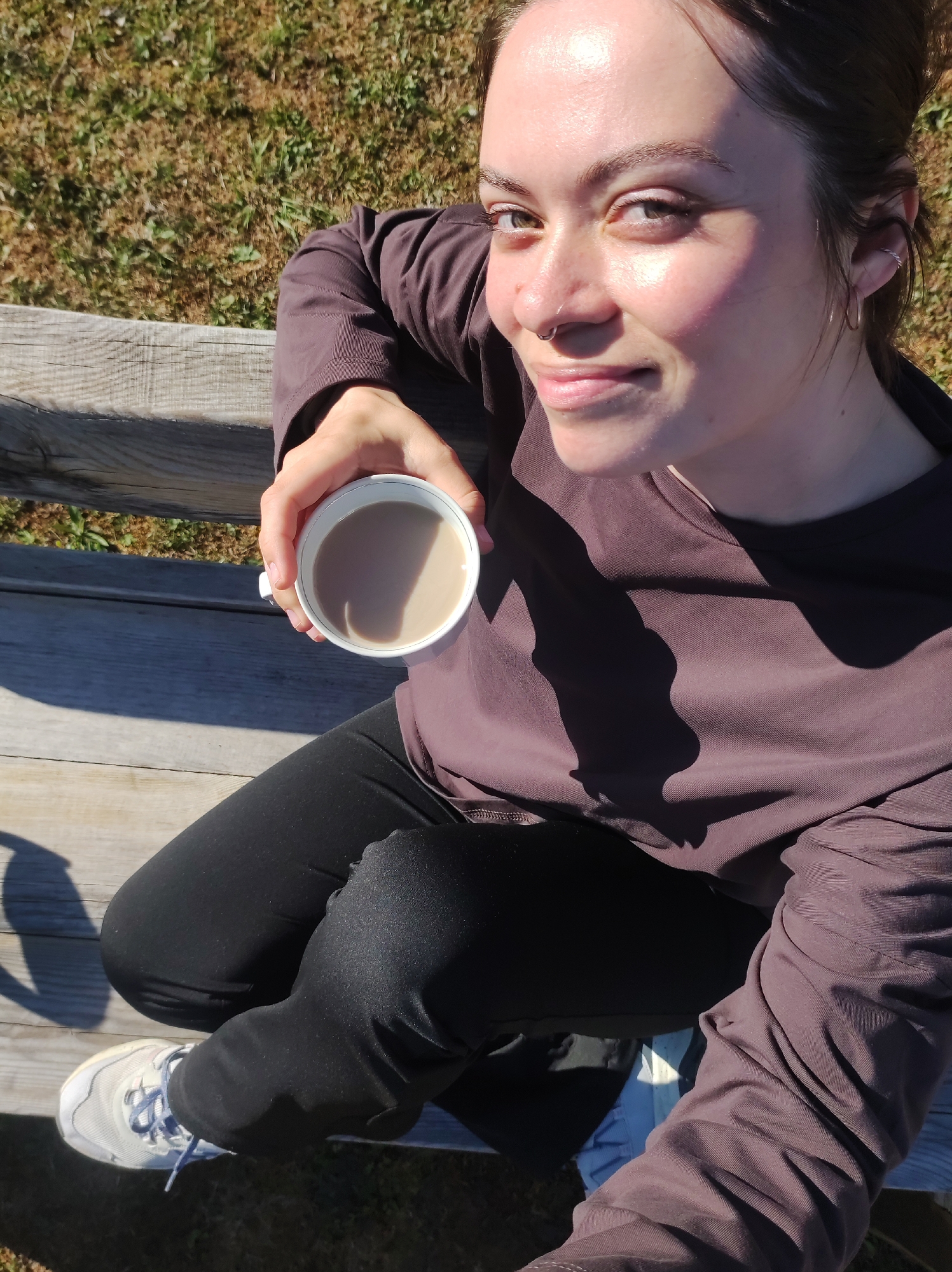A woman is sitting on a weathered wooden bench outdoors, holding a white cup filled with a light brown liquid, likely coffee. She is wearing a long-sleeved brown shirt and black leggings, with white sneakers with blue laces on her feet. The scene is bathed in sunlight, creating stark shadows on her clothing and the bench. The background shows grassy ground. No specific landmarks visible to pinpoint the location in the city of Фоки, Russia.