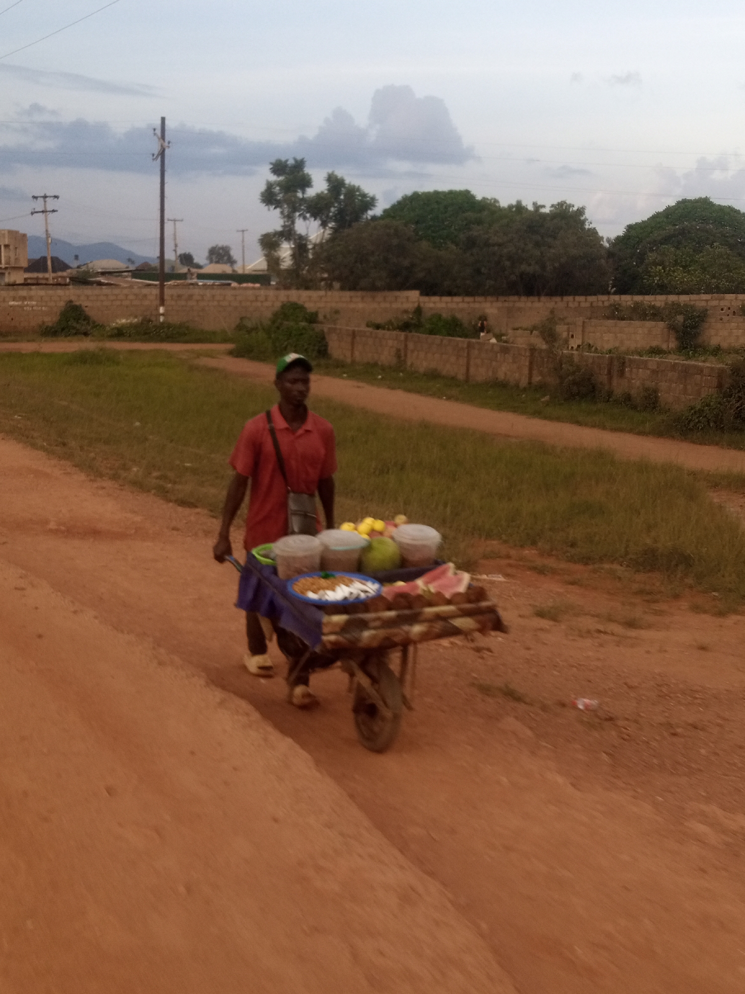 A man is pushing a wheelbarrow full of goods down a dirt road in Kwang, Nigeria. The man is wearing a red short-sleeved shirt and a green baseball cap. The wheelbarrow is covered with a blue tarp, and it is filled with various items, including fruits like watermelon, and containers of what appear to be nuts or snacks. The road is dirt, and there is grass and some foliage on either side. In the background, there are brick walls, trees, and some buildings. A utility pole with wires is also visible. The sky is overcast, suggesting it might be late afternoon or evening. The overall scene depicts a vendor on the move, likely selling goods. There is no visible text in the image.