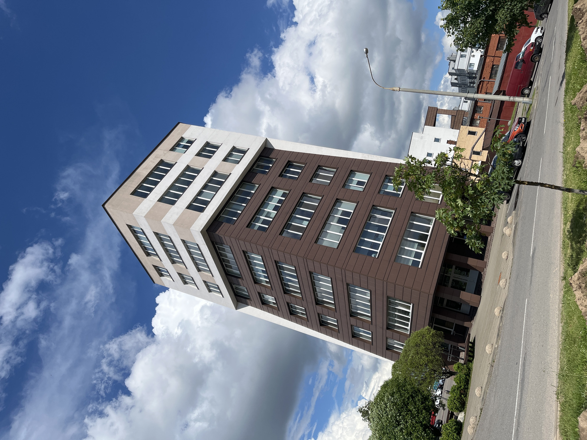 The image shows a modern apartment building with a slanted facade, located on a street in Minsk, Belarus. The building is tall and narrow, with alternating bands of brown and beige on its exterior. It has numerous windows on each floor, reflecting the bright blue sky and white clouds. In the foreground, there is a paved sidewalk with decorative stone bollards, a grassy area, and a road. Several cars are parked along the street, including a red van and an orange car. Trees and bushes line the sidewalk and the edges of the property. Other buildings, including some brick structures and modern residential complexes, can be seen in the background. A streetlamp is visible on the right side of the image. The overall scene depicts a typical urban environment on a sunny day.