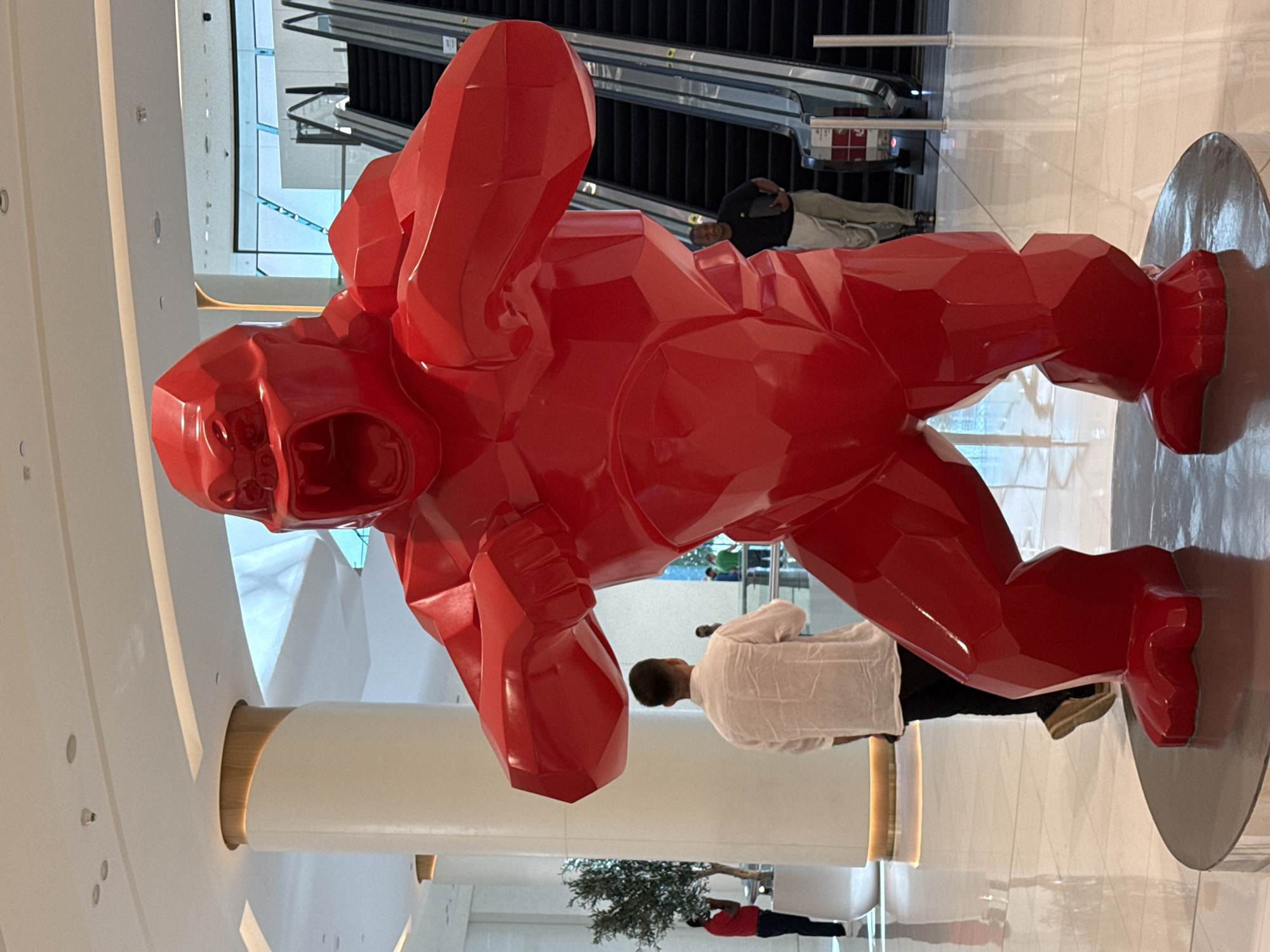 A large, red, polygonal sculpture of a roaring gorilla stands prominently in the foreground of a modern, well-lit interior space. The sculpture has sharp angles and geometric facets, giving it a stylized, almost crystalline appearance. The gorilla's mouth is open in a roar, its teeth bared.
In the mid-ground, a man wearing a white, long-sleeved shirt and dark pants stands with his back to the camera, looking towards the sculpture. To the left, a potted tree with sparse green foliage is visible next to a tall, cylindrical white pillar with a wooden accent. Further in the background, a set of escalators is visible, with people ascending and descending. One person in a dark t-shirt and light-colored pants is seen near the escalators, holding a black tablet or folder. Another person in a red shirt is visible to the far left, near the potted tree.
The floor is highly reflective, indicating a polished surface, possibly marble or tile, mirroring the overhead lighting and the sculpture. The overall environment suggests a large public space such as a shopping mall or an airport terminal. The scene is likely located within Dubai, United Arab Emirates, a city known for its modern architecture and large-scale public art installations. The lighting is bright and diffuse, illuminating the space evenly.