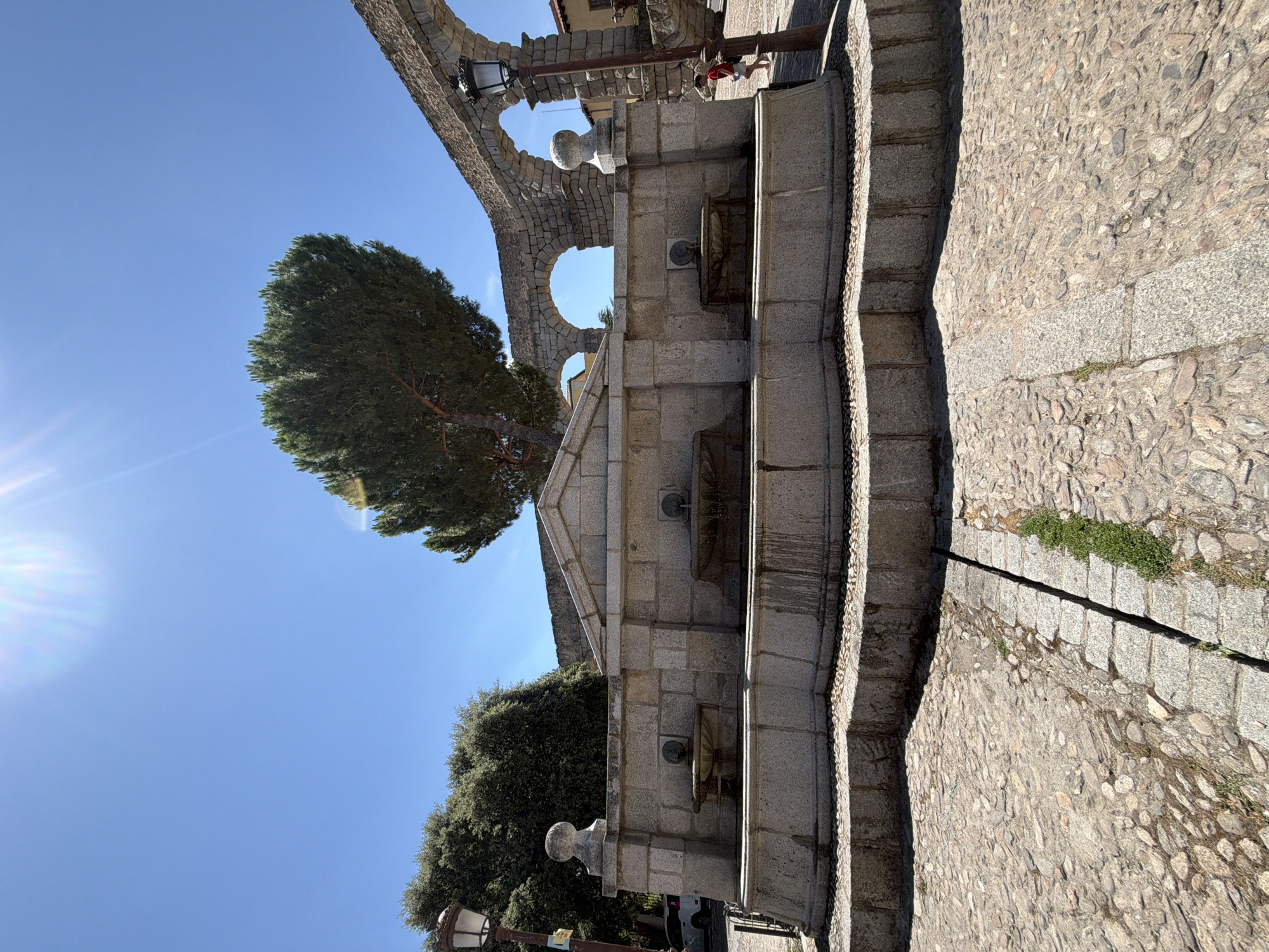 This is a full shot image of the Fountain of the Three Cultures (Fuente de los Tres CUlturas) in Segovia, Spain. The fountain is made of granite blocks, with three basins. Above the basins is a triangular pediment. In the background, a large pine tree stands in front of a section of the Roman Aqueduct of Segovia. The ground in front of the fountain is paved with cobblestones. A lamppost is visible to the left of the fountain. A person wearing a red shirt and white shorts is walking past the fountain on the right side of the image, moving away from the camera. The sky is clear and blue, with the sun creating lens flare in the upper left corner.