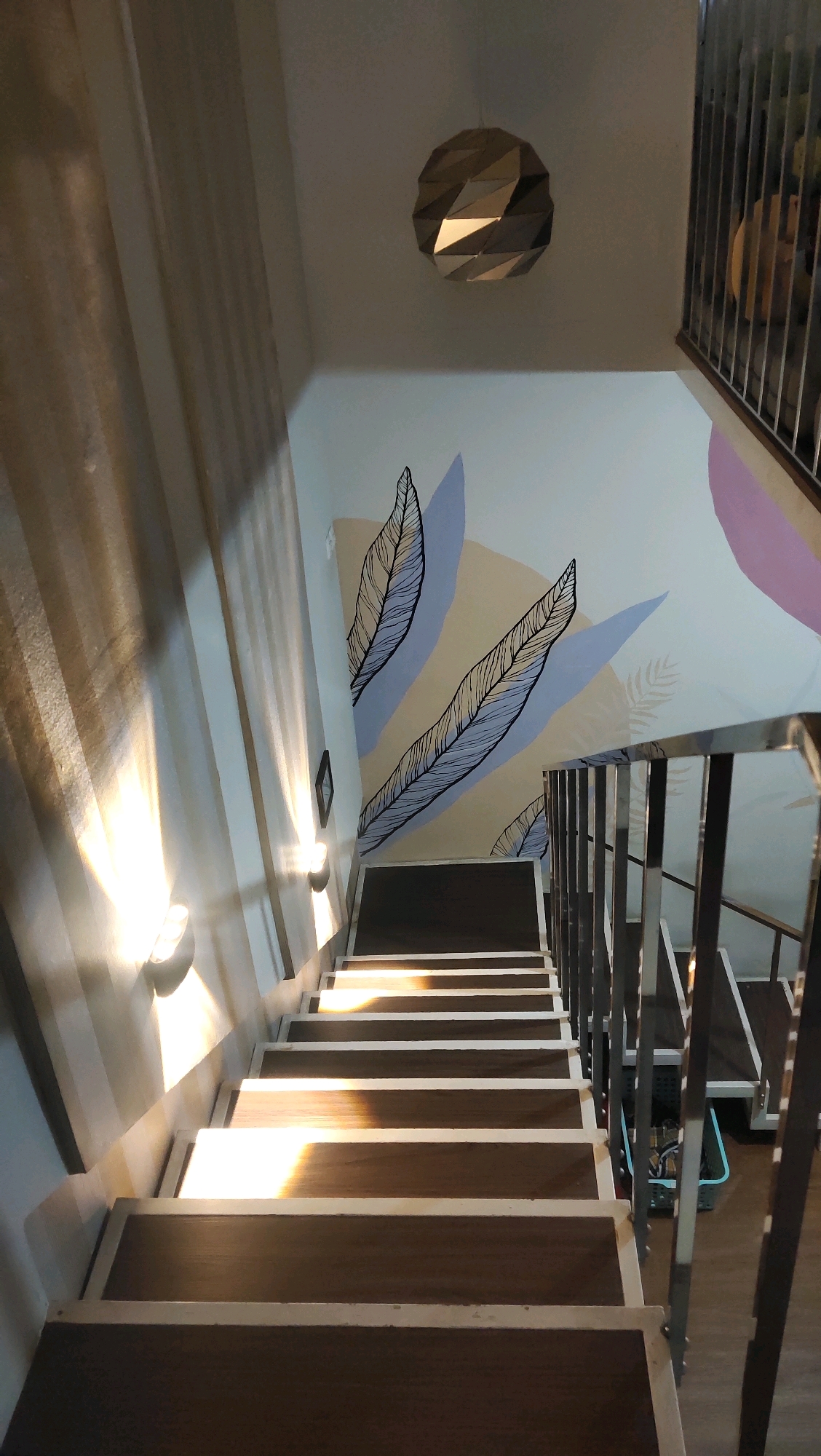 The image shows a dimly lit interior staircase with wooden steps. Sunlight streams in from the left, casting diagonal shadows across the stairs and illuminating the wall on the left. The wall opposite the sunlight is adorned with a mural of large, stylized leaves in shades of light purple, beige, and pink. A geometric pendant light fixture hangs from the ceiling. A metal railing runs along the right side of the staircase. The overall impression is one of a modern, indoor space with artistic decoration. The location is identified as Karangwangkal, Indonesia.