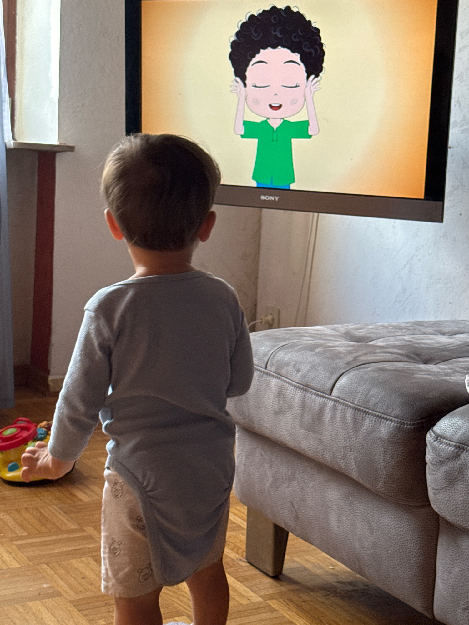 A young child, facing away from the camera, stands on a wooden floor in a room, watching a television screen. The child is wearing a light grey long-sleeved shirt and a diaper with a pattern of small bear faces. To the left of the child is a colorful toy, partially visible. The television screen displays a cartoon character with curly black hair, closed eyes, and hands near its temples, wearing a green shirt and blue shorts. A large, tufted grey sofa occupies the right side of the frame. The background consists of a white wall with some textural variation and a window with a light blue curtain on the left. The room appears to be a living space. Based on the presence of the television and sofa, and the general domestic setting, this scene could be located within a residential property in the city of Überherrn, Germany.
