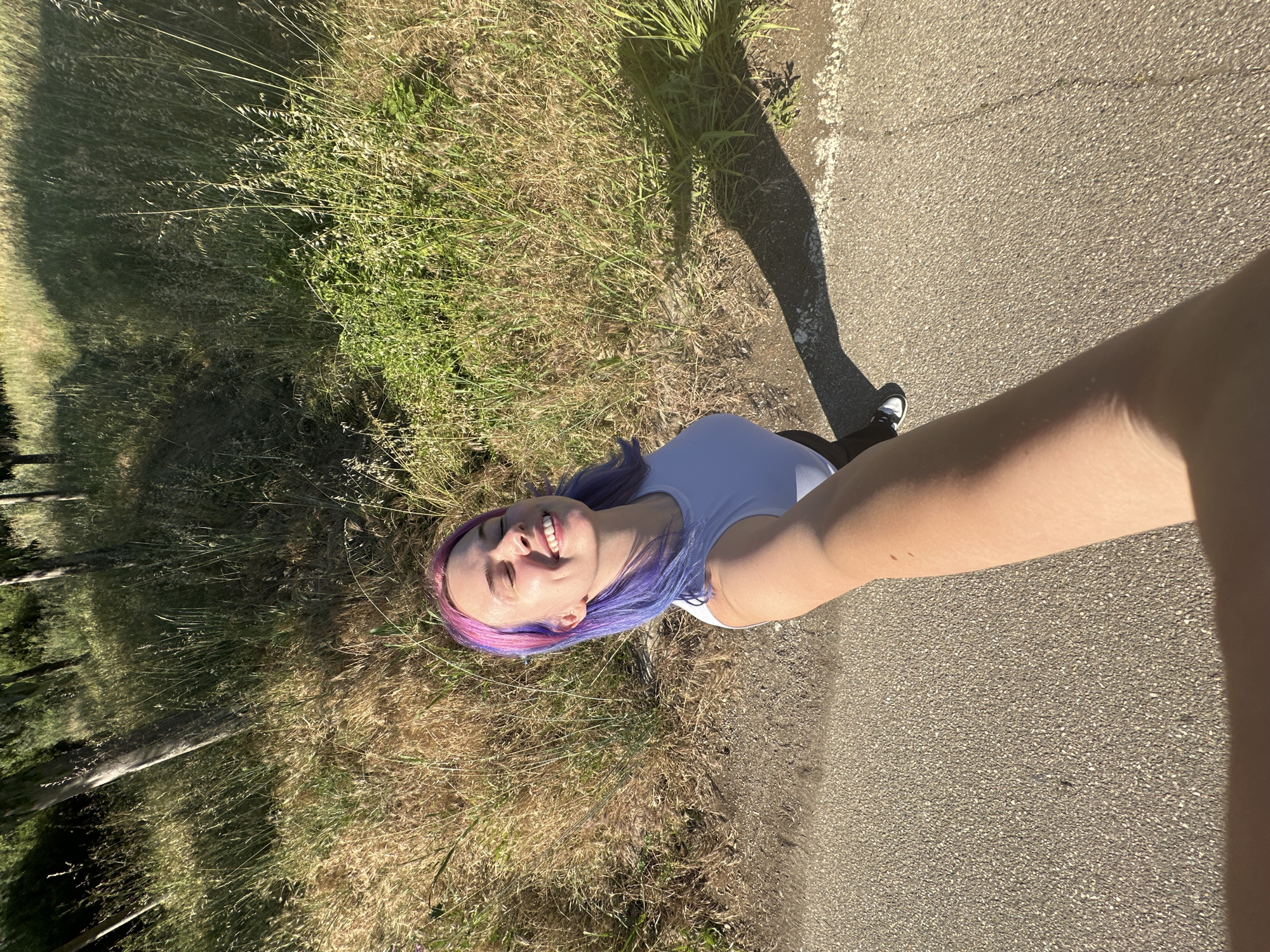 A medium-shot, eye-level photograph shows a woman with purple and pink hair smiling and looking up at the camera. The photograph appears to be a selfie, taken with the woman's arm extended. She is wearing a white shirt. The background includes tall, dried grass and a paved path. The lighting suggests a sunny day. The location is within Caissargues, France.