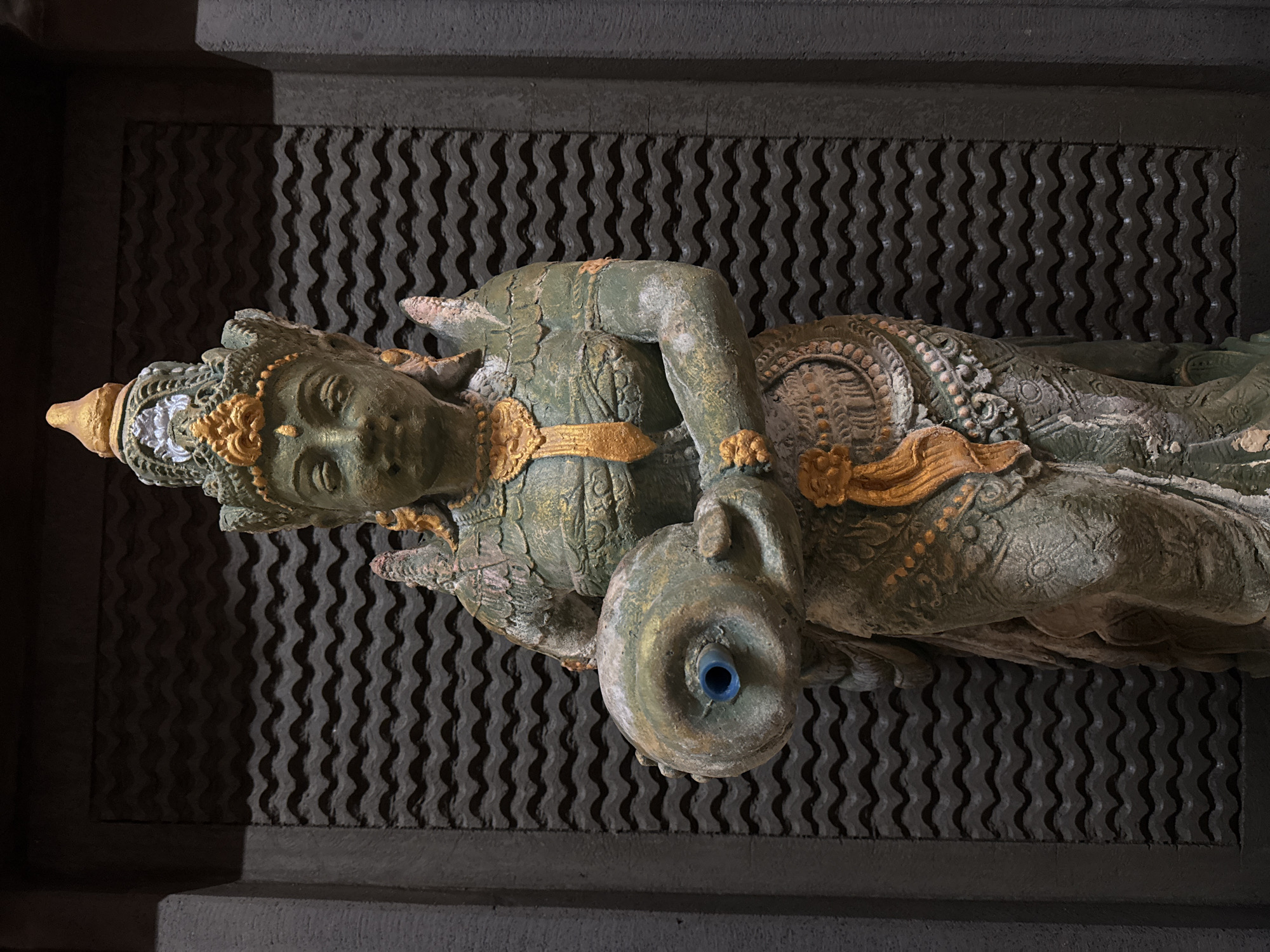 This media file features a detailed statue, likely made of stone or a similar material, with a weathered, aged appearance. The statue is predominantly a muted green color, with accents of gold and some areas of lighter, pinkish-grey wear. It depicts a figure with intricate headdresses, jewelry, and ornate clothing. The figure holds a round object, possibly a vessel or pot, which has a blue insert in its center. The statue is oriented vertically in the frame.
The backdrop behind the statue is a textured, dark grey surface with a repeating wavy pattern. Above and below this textured surface are darker, flat surfaces, suggesting it's part of a wall or architectural feature. The lighting creates strong shadows, particularly on the wavy background, emphasizing the texture.
The scene appears to be indoors or in a shaded area, as there is no indication of natural daylight or weather conditions. The overall aesthetic suggests a cultural or historical context, possibly a religious or decorative element in a building. Given the provided location context of Kuta, Indonesia, the statue likely represents a figure from Balinese or Indonesian mythology or art. There is no visible text in the image.