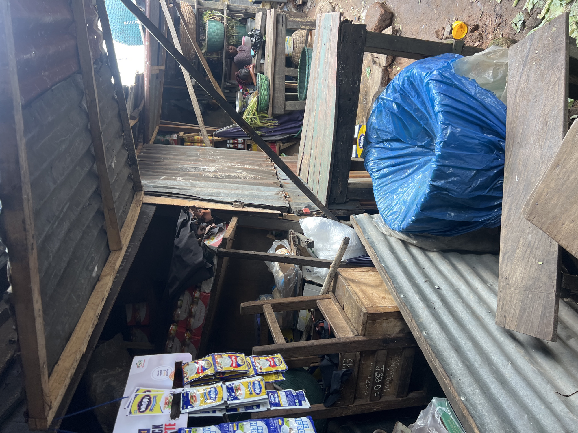 This image depicts a bustling market stall in Nkpor, Nigeria. The stall is constructed from corrugated metal and wood, with a corrugated metal roof. Goods are displayed on shelves and hanging racks. Items visible include packaged food products, baskets of produce, and various other items associated with a market setting. In the background, two women are visible behind market tables, one appearing to be attending to produce and the other seated. The ground surface is a mixture of dirt and rough stones. The overall impression is one of a vibrant, active marketplace.