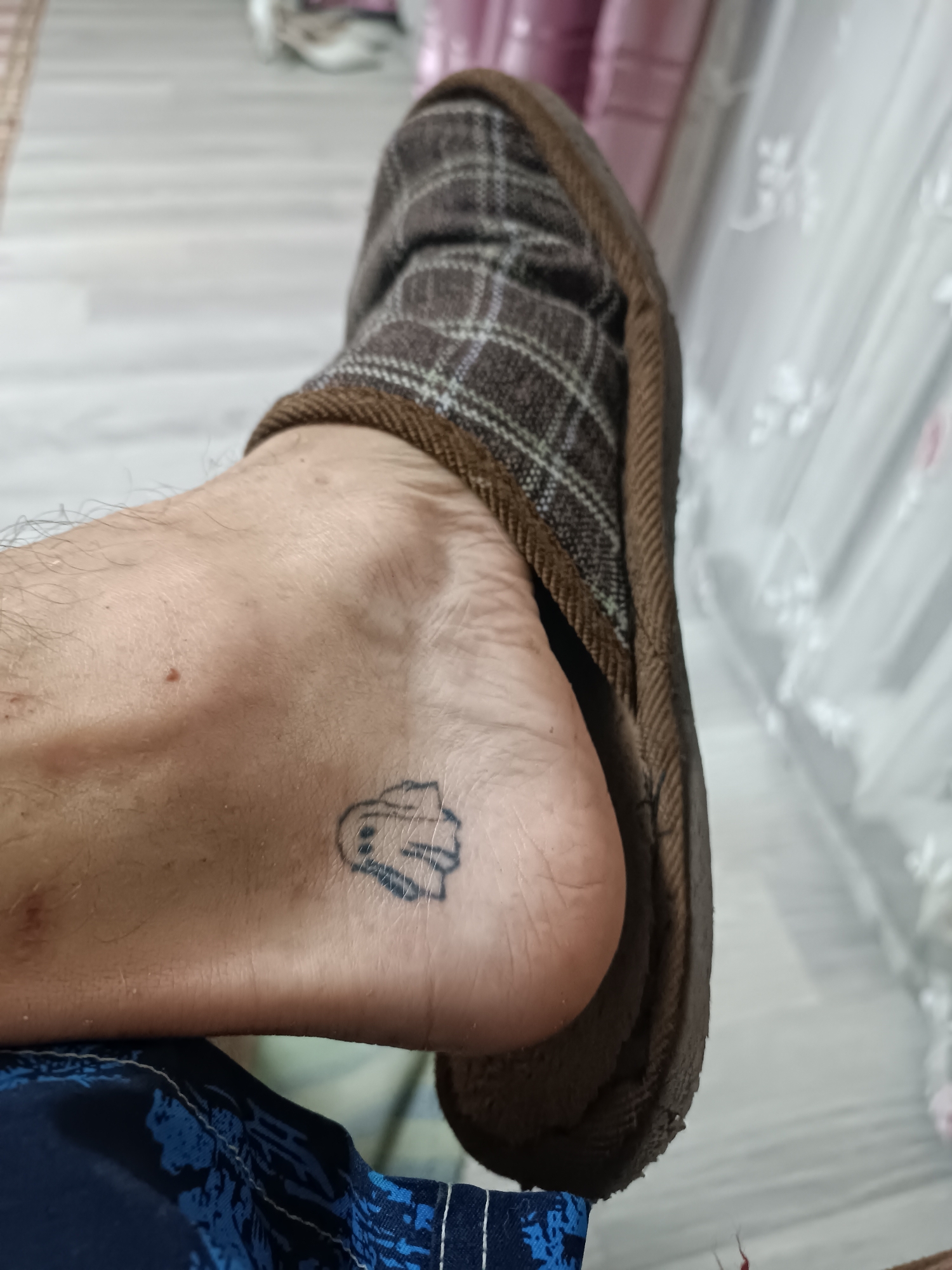 A close-up, eye-level, medium shot focuses on a person's bare foot, which has a small, dark blue tattoo of a simplified cartoon ghost on its inner arch. The foot is positioned with the heel towards the bottom right of the frame. A brown and grey plaid slipper is partially visible, covering the top and back of the foot. The surrounding environment is blurred but appears to be an indoor setting with a light-colored floor and white curtains or blinds in the background. The person is wearing dark blue patterned shorts.