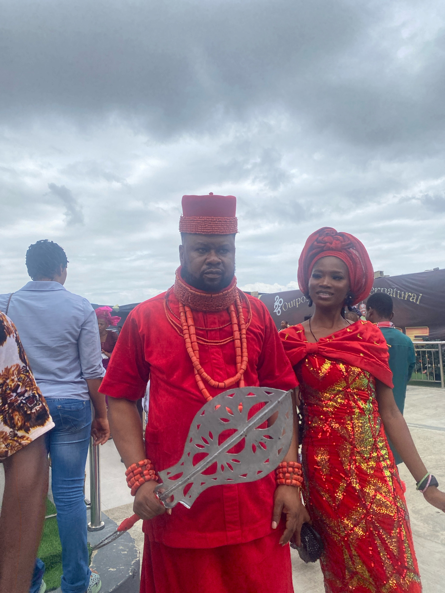 The image shows a couple in traditional red attire, likely at an outdoor event in Ado, Nigeria. The man on the left wears a red cap, a red beaded necklace, and a red outfit, holding a silver, ceremonial staff. The woman on the right is adorned in a red headwrap and a red sequined dress. Behind them, other attendees and a banner that reads "SUPERNTURAL" are visible. The sky is overcast with clouds.