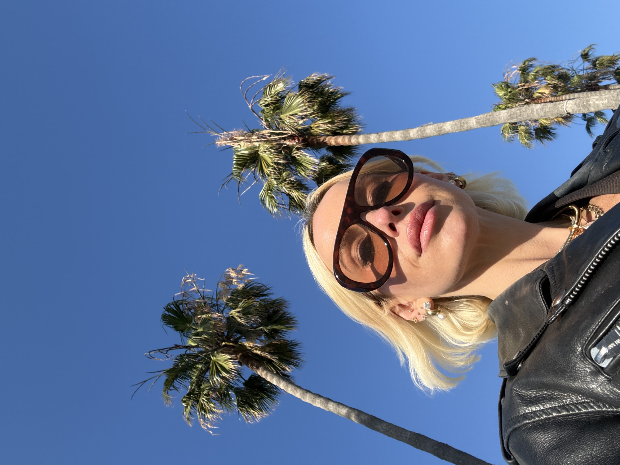 A low-angle, close-up photograph shows a person with blonde hair and sunglasses, against a background of palm trees and a clear blue sky. The person is wearing a black leather jacket and gold jewelry. The location is likely Los Angeles, United States of America, given the palm trees and clear sky, a common feature of this area.