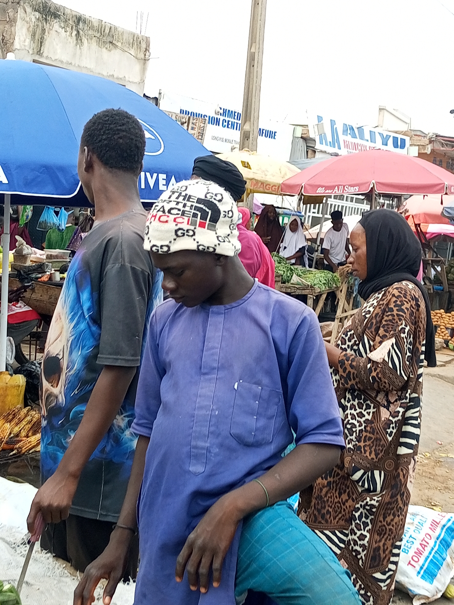 In Tempure, Nigeria, a street market scene unfolds under a cloudy sky. A person in a blue top with a graphic print on the back is standing, looking towards the right. Beside them, a young boy in a light purple traditional outfit is looking down. To the right, a woman wearing a leopard-print dress and a black headscarf is facing the left, with a market stall of produce behind her. Various umbrellas, including a large blue one and a red one, provide shade over the market stalls. In the background, buildings and signage are visible, with one sign partially obscured by an umbrella and reading "AHMED PROVISION CENTER." A white sack with red and blue lettering, reading "BEST QUALI TOMATO MILL," is on the ground to the right. The ground is dusty and appears to be unpaved. The overall atmosphere suggests a busy marketplace.
