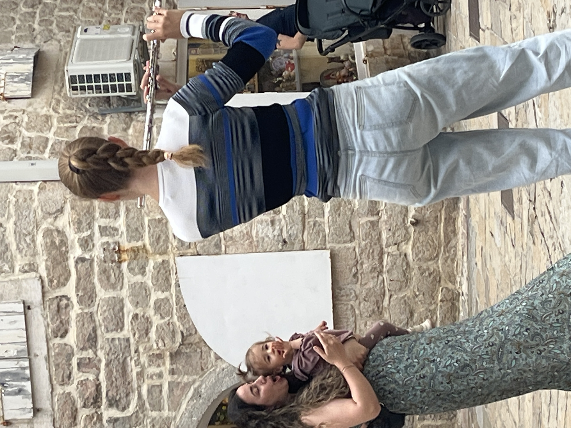An outdoor scene shows a woman playing a flute while standing near a stone wall. She has blonde hair in a braid, wears a striped shirt, and light-colored pants. Behind her, a black stroller is visible, along with an air conditioning unit mounted on the wall. In the lower part of the image, another woman is seen holding a baby in her arms. They are positioned in front of a large, arched opening in the wall.
Considering the architectural details, specifically the stone wall and arched opening, this image is very likely captured in the Old Town of Budva, Montenegro. The architecture is very typical of the region.