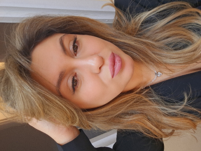 A close-up portrait depicts an adult female indoors, located in Protasovo, Russia. The woman has long, wavy, blonde to light brown hair with highlights, styled with significant volume. Her right hand is partially visible in the upper left quadrant of the frame, positioned within her hair. Her face displays dark eyes with visible eyeliner and long eyelashes, groomed eyebrows, and full lips colored with pink lipstick. Her gaze is directed slightly downwards and to the viewer's right. She is wearing a dark, V-neck garment. Around her neck is a necklace featuring a thin, light-colored chain and a small, intricate pendant, possibly heart-shaped and adorned with multiple clear stones. The background is out of focus, indicating an indoor setting with indistinct light and dark elements.
