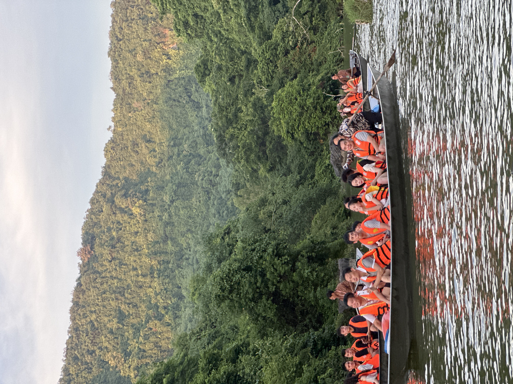 In the image, a group of people are on a boat ride on a river. The people are wearing orange life jackets. The boat is moving through the water and the people appear to be enjoying the scenery. In the background, there are lush green trees and mountains. The sky is partly cloudy. The scene suggests a tourist activity in a natural setting. The location is in the city of Yên Sơn Ward, Vietnam.