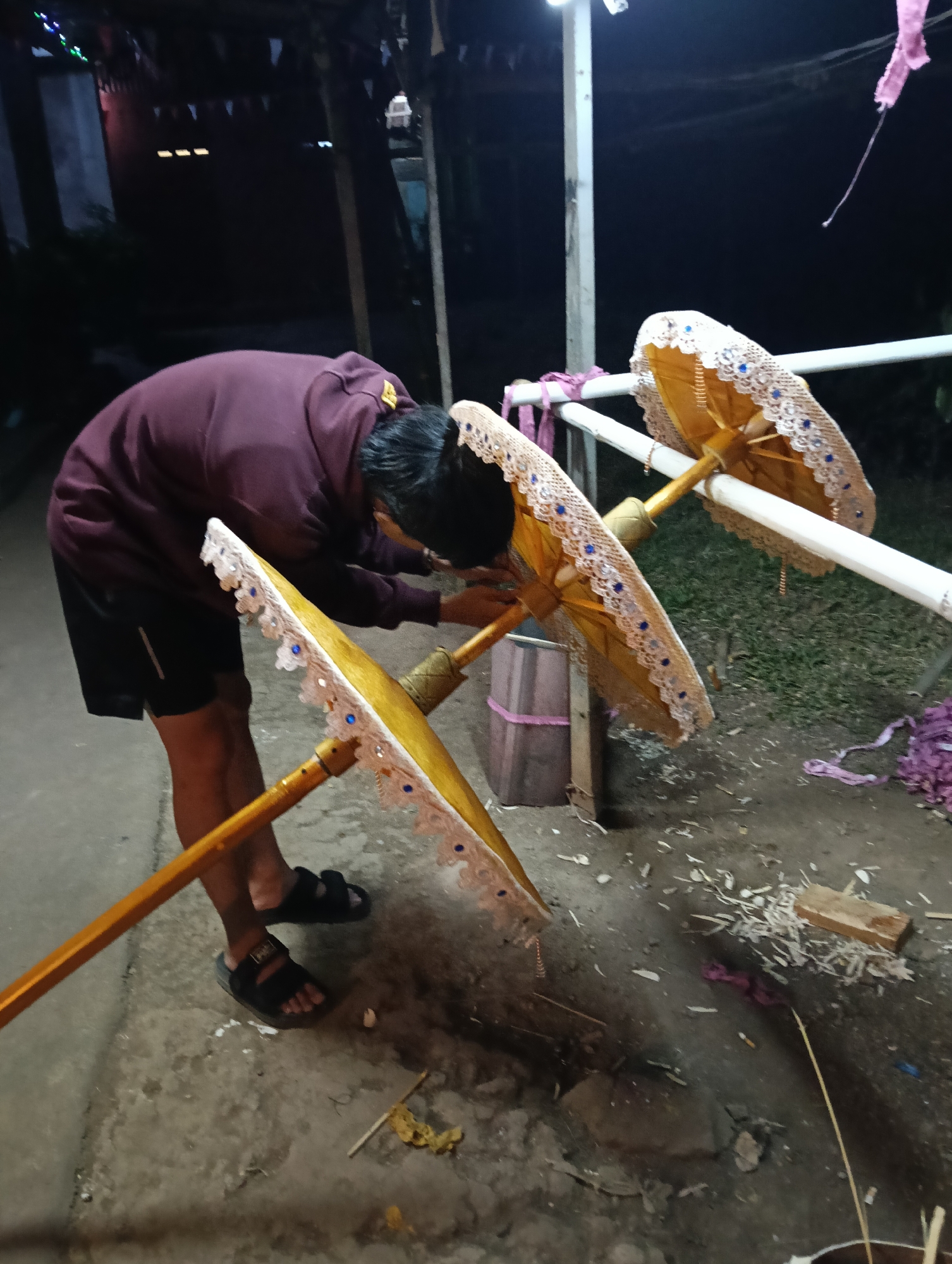 In Tasikmalaya, Indonesia, a person is seen working on decorative umbrellas outdoors at night. The individual, wearing a maroon hoodie and black shorts, is focused on one of the umbrellas. The umbrellas are ornate, featuring gold fabric and delicate white lace trim with blue embellishments. They are mounted on long wooden poles and are propped up by a white railing. Scattered debris, possibly wood shavings and fabric scraps, are visible on the ground around the work area, suggesting a crafting or preparation process. The background is dark, illuminated by artificial lighting, and hints at an outdoor, possibly semi-rural, setting.