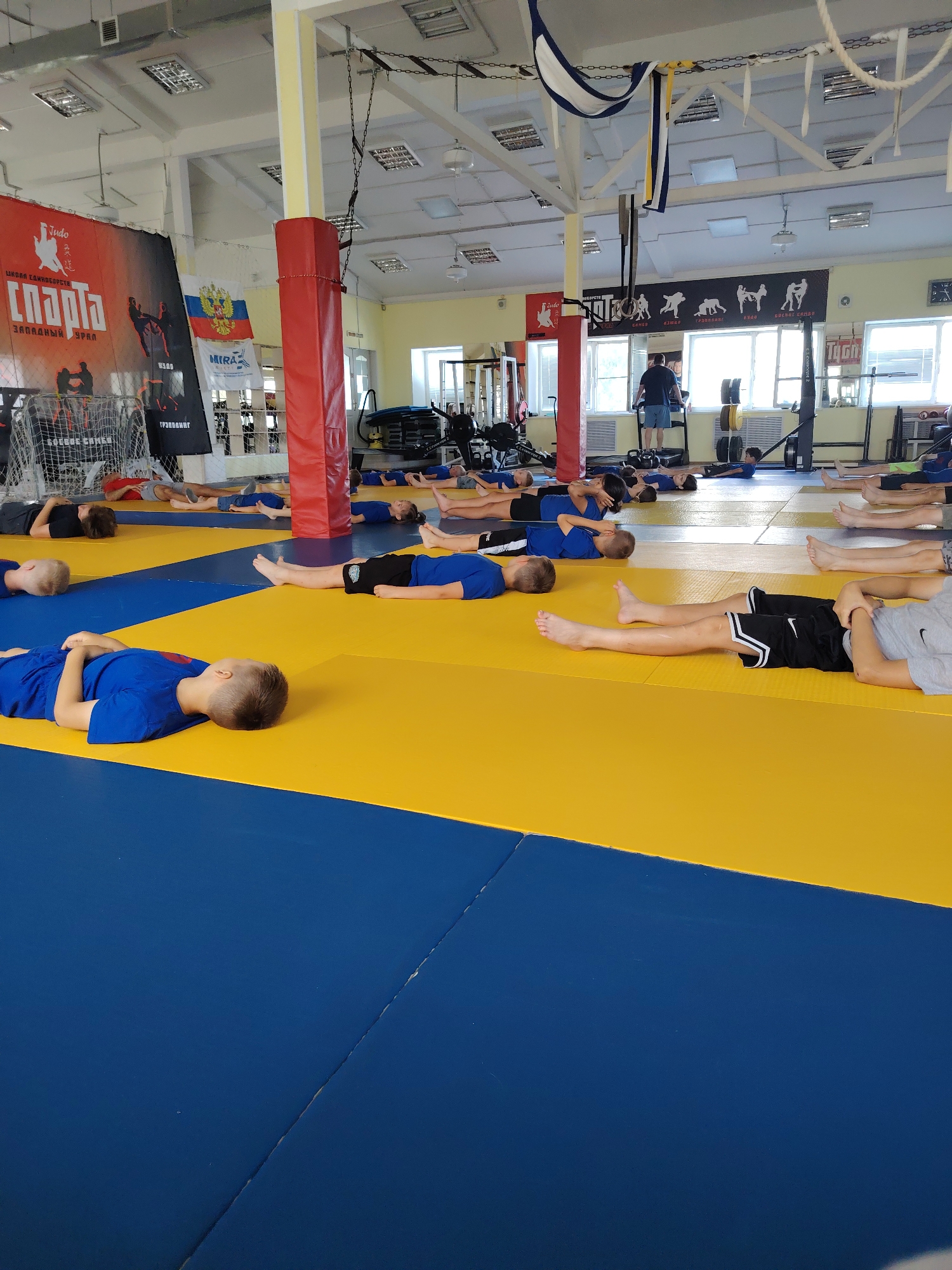 This is a full shot of a dojo in Chaikovsky, Russia, where a group of children are lying on a blue and yellow mat. The children are mostly in blue uniforms and appear to be engaged in some sort of exercise or rest period. They are positioned in rows, facing different directions. In the background, there are various pieces of sports equipment, including weightlifting machines, a treadmill, and a punching bag. Banners and flags are also visible on the walls, indicating the location is a sports training facility. One banner displays the word "SPARTA" in large letters, along with images of martial arts. The overall environment suggests a well-equipped and active training center.