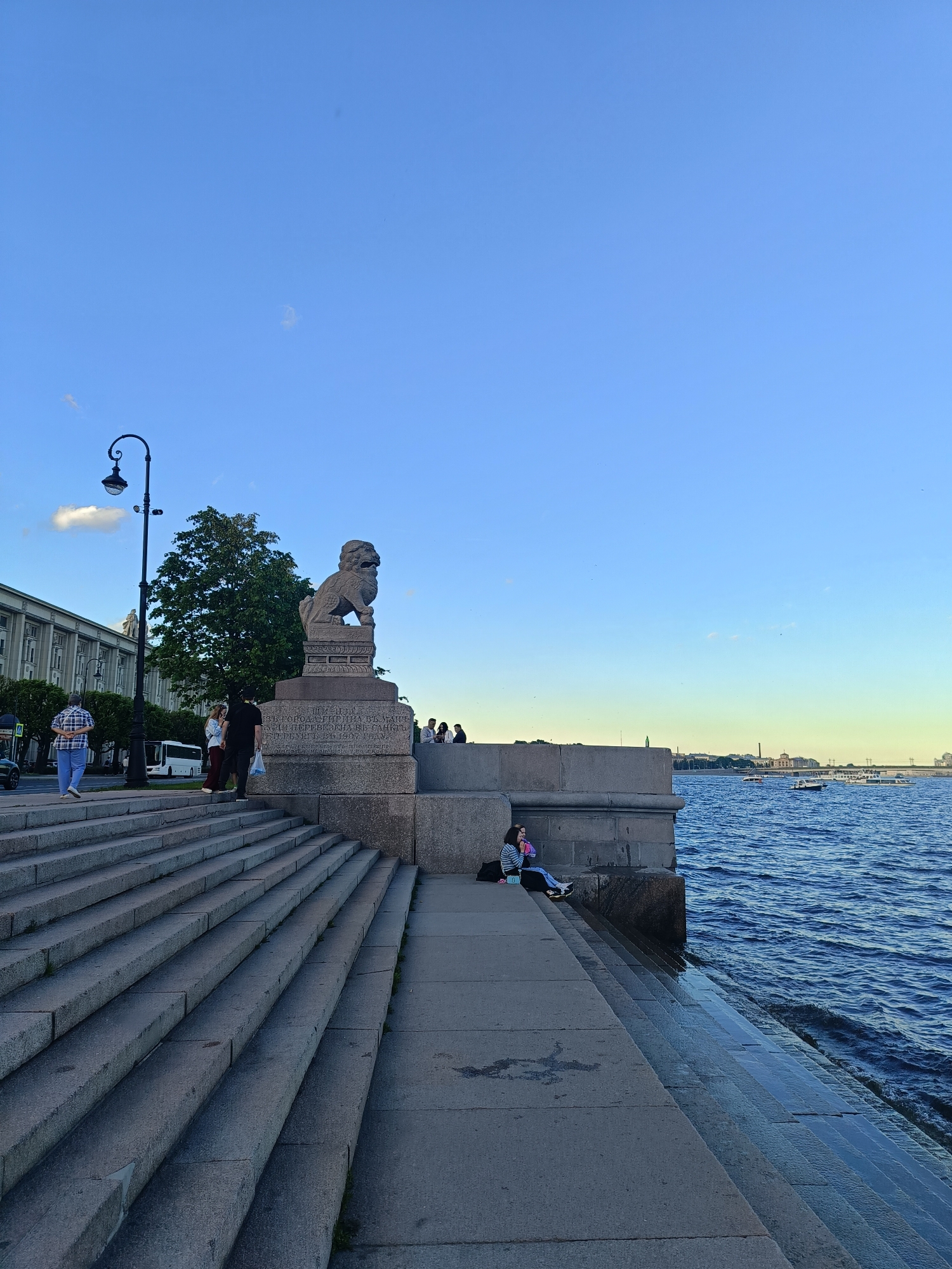 A wide shot captures a waterfront scene in Saint Petersburg, Russia, under a clear blue sky with a hint of clouds. In the foreground, a grand stone staircase descends towards a dark body of water, likely the Neva River, which is choppy with ripples. On the left side of the image, a stately building with multiple windows and an arched entrance stands behind a sidewalk and a lamppost. Several people are visible on the sidewalk and on the stairs.
To the right of the stairs, a large stone pedestal supports a statue of a lion, its head tilted forward. The pedestal is inscribed with Russian text. A woman is seated on the edge of the stone embankment near the lion statue, looking towards the water. In the distance, the river is dotted with several boats. The architecture and the presence of the lion statue suggest this location is near the Peter and Paul Fortress or the St. Petersburg State University.