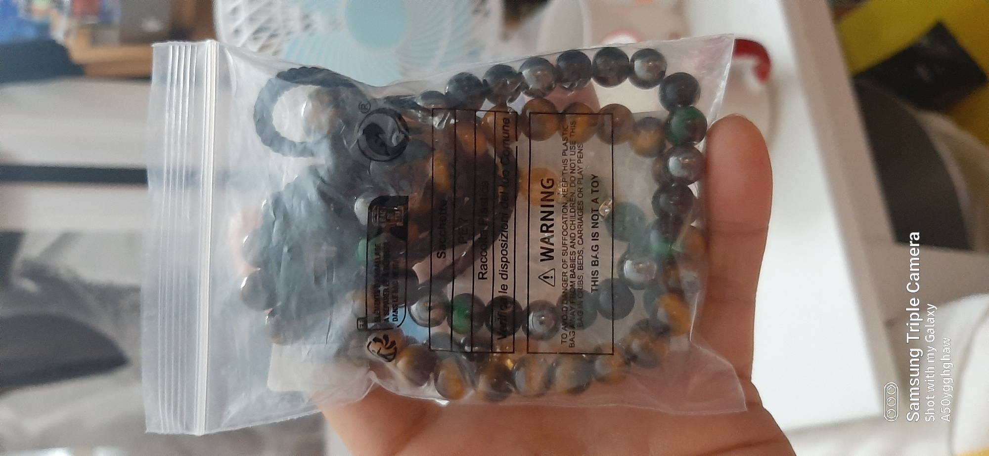 The image shows a close-up of a person's hand holding a clear plastic bag containing bracelets. The bracelets are made of round beads in various earth tones, including brown, amber, and green. A dark-colored charm or pendant is attached to the bracelets. The plastic bag has text printed on it, including a "WARNING" label indicating the danger of suffocation from the bag for babies and children, and stating that the bag is not a toy. The background is blurred but suggests an indoor setting with what appears to be a fan and some other items on a surface. The lighting is bright, indicating daytime. The image is accompanied by text indicating it was taken with a Samsung Triple Camera on a Galaxy phone.