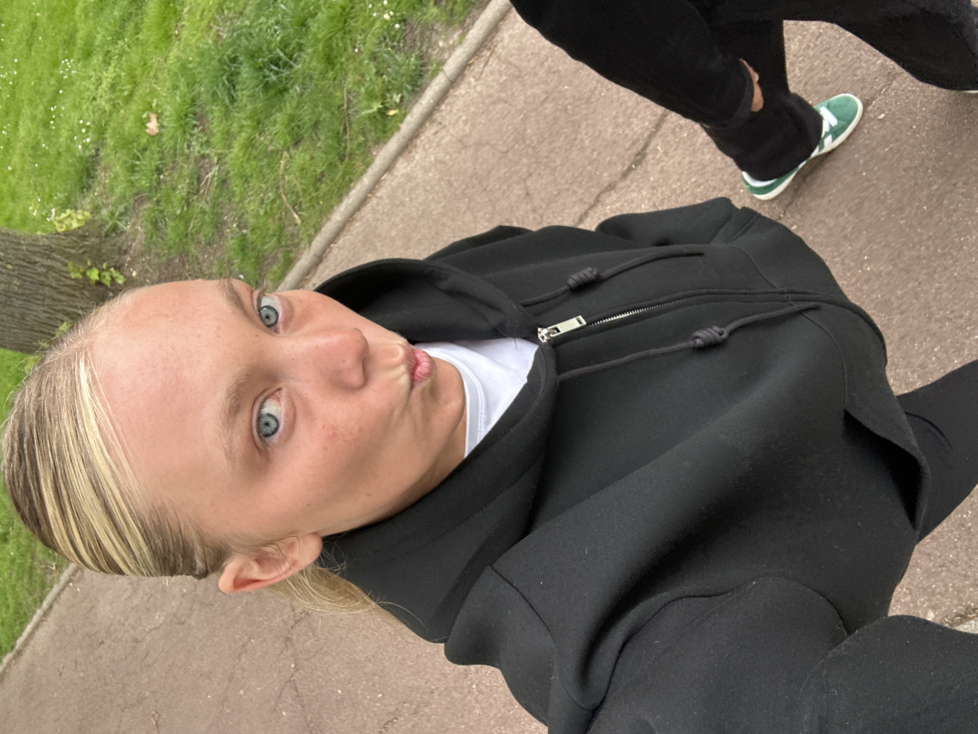 A young, light-skinned woman with blond hair and blue eyes is taking a selfie outdoors. She is wearing a white t-shirt under a black zip-up hoodie and black pants. She has puckered lips. Another person's legs and green sneakers are partially visible in the upper right corner. The ground is paved, and there is grass and a tree visible in the upper left corner. Based on known imagery, the scene could plausibly be located in Noisiel, France.