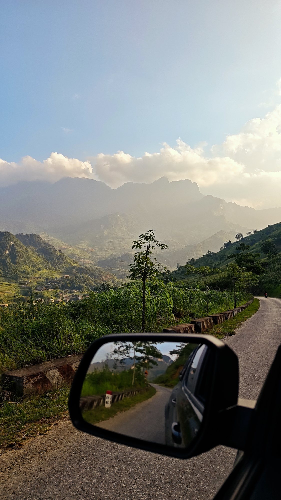 The image depicts a scenic view of the mountainous landscape near Du Già Commune, Vietnam, observed from within a vehicle. The foreground features the side mirror of a car, reflecting a stretch of asphalt road curving through the terrain. Tall green vegetation, including grass and small trees, lines the roadside. In the distance, prominent mountains rise, partially shrouded in mist and illuminated by the sun. A few scattered buildings of a rural settlement are nestled in the valleys between the slopes. The sky is partly blue with scattered clouds. A motorbike can be seen on the road in the distance.