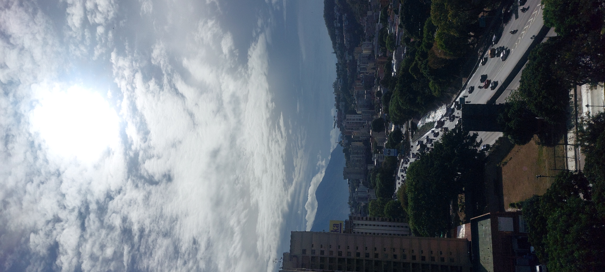This media file offers an elevated, panoramic view of a vibrant city, likely Caracas, Venezuela, illuminated by a bright, daytime sky. The upper portion of the scene is dominated by a brilliant sun glare, surrounded by scattered white, cumulus clouds set against a pale blue sky.
In the background, a majestic mountain range stretches across the horizon, its base partially veiled by clouds, characteristic of the El Ávila mountain range that overlooks Caracas. The midground and foreground reveal a densely populated urban area, featuring numerous multi-story buildings of varying architecture, suggesting a mix of residential and commercial properties.
A wide, multi-lane highway or major road cuts through the urban sprawl, bustling with moderate to heavy vehicle traffic. Many cars are visible on the road, indicating active movement and daily commuting. Lush green trees line the highway and are interspersed among the buildings, adding pockets of nature to the cityscape. A section of dry, brown land can be seen adjacent to the road in the foreground.
The intense brightness of the sun indicates that the photograph was taken during midday or early afternoon. The weather appears fair, with bright sunshine breaking through the clouds. A notable detail is a yellow sign with blue lettering on the roof of a building in the midground, which appears to read "BNC," a common abbreviation for Banco Nacional de Crédito, a bank found in Venezuela, further confirming the location context. No individual people are clearly discernible. The overall scene captures the energetic rhythm of an urban center set against a dramatic natural mountainous backdrop.