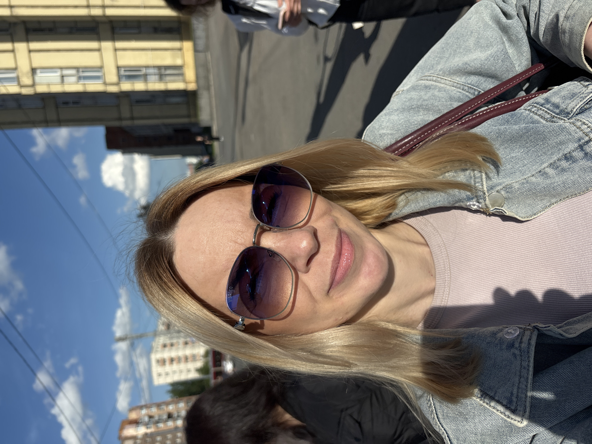 A bright outdoor portrait captures a woman with blonde hair and fair skin, wearing sunglasses, looking upwards with a faint smile. She is dressed in a light-wash denim jacket over a light pink or mauve ribbed top. A maroon-colored strap, possibly from a bag, rests on her shoulder. The sunglasses have silver frames and reflective purple-blue lenses, with the brand name "TRUSSARDI" visible on the left lens frame.
The setting appears to be an urban environment on a sunny day. The sky is a clear blue with scattered white clouds, reflected prominently in the woman's sunglasses. In the background, various buildings are visible, including a multi-story yellow building with multiple windows and other residential-style structures. Overhead power lines crisscross the sky. The ground below is a paved surface, likely asphalt, with long, distinct shadows of people indicating strong sunlight, suggesting it is mid-day or early afternoon. Shadows of other individuals are cast on the ground, and parts of other people, including hands holding a phone, are subtly visible in the background, implying a public outdoor space or street. The overall impression is one of a relaxed moment captured in a bustling, sunlit city. The image is rotated 90 degrees clockwise from standard orientation.